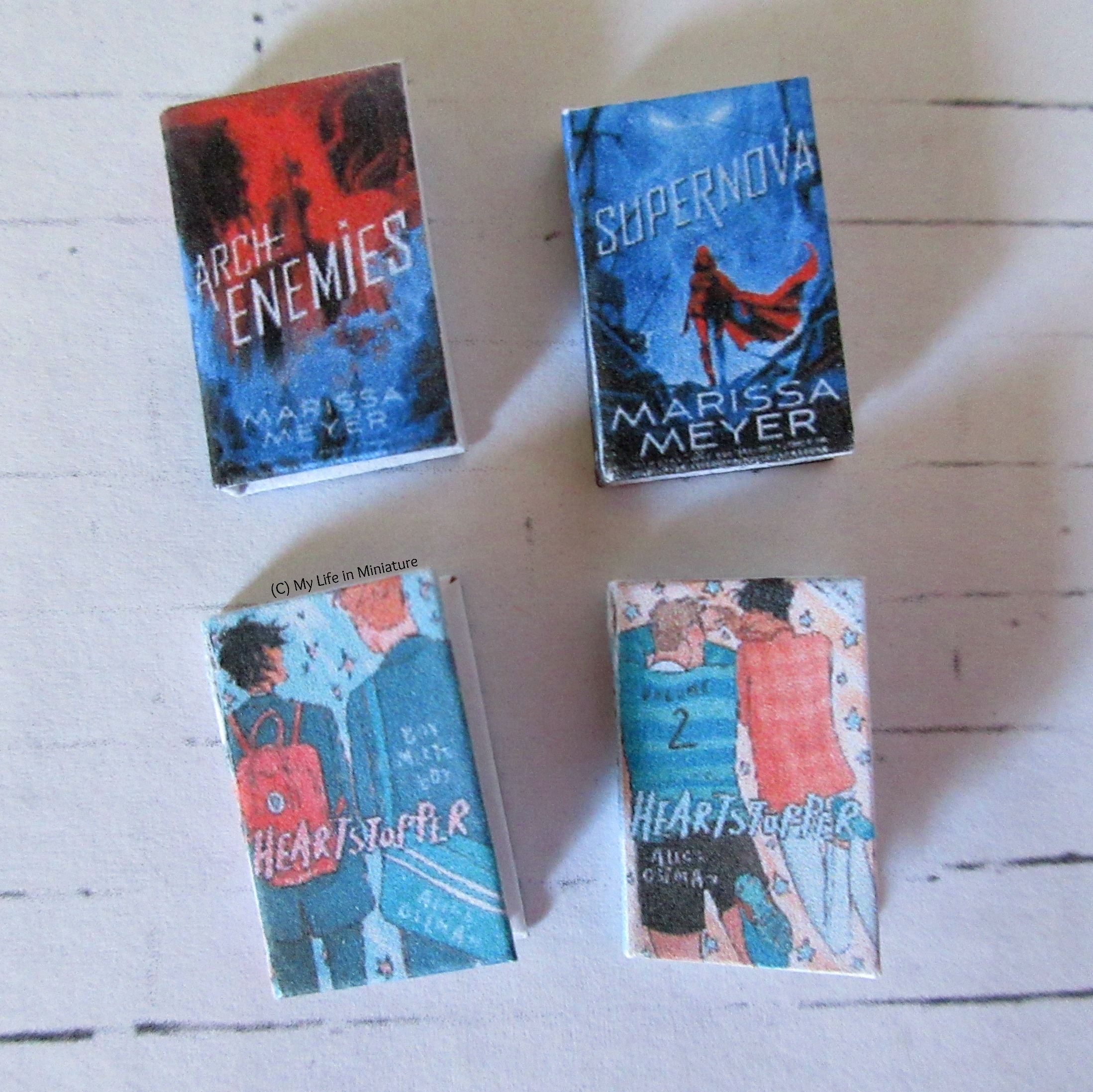 Four miniature books lie on a white brick background, covers facing the camera. Archenemies and Supernova by Marissa Meyer are above Heartstopper volumes 1 and 2 by Alice Oseman.