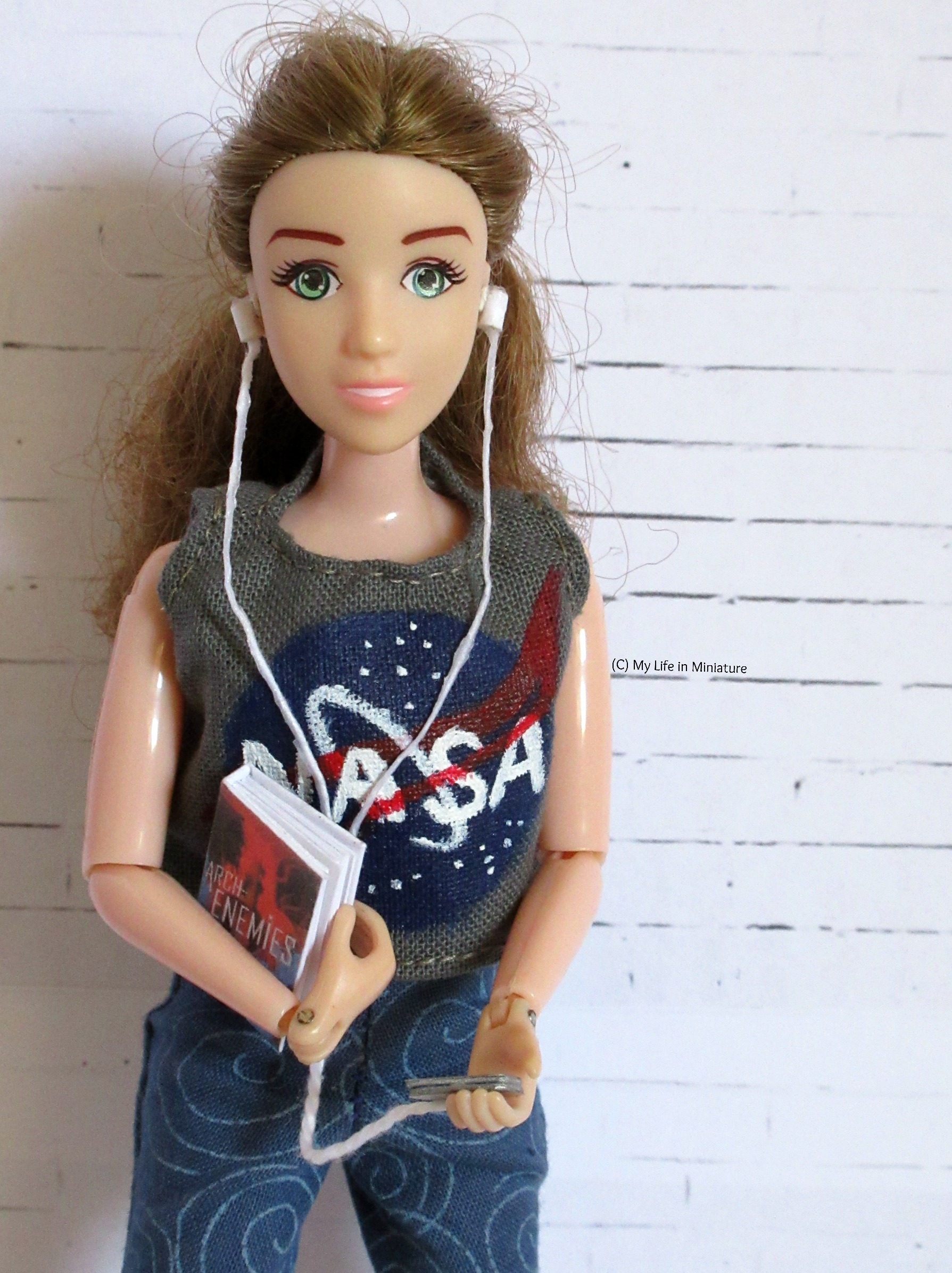 Sarah faces the camera against a white brick background. She wears a dark grey NASA t-shirt, and carries her phone in her hand. Connected to her phone is a set of wired earbuds, like the older Apple ones.