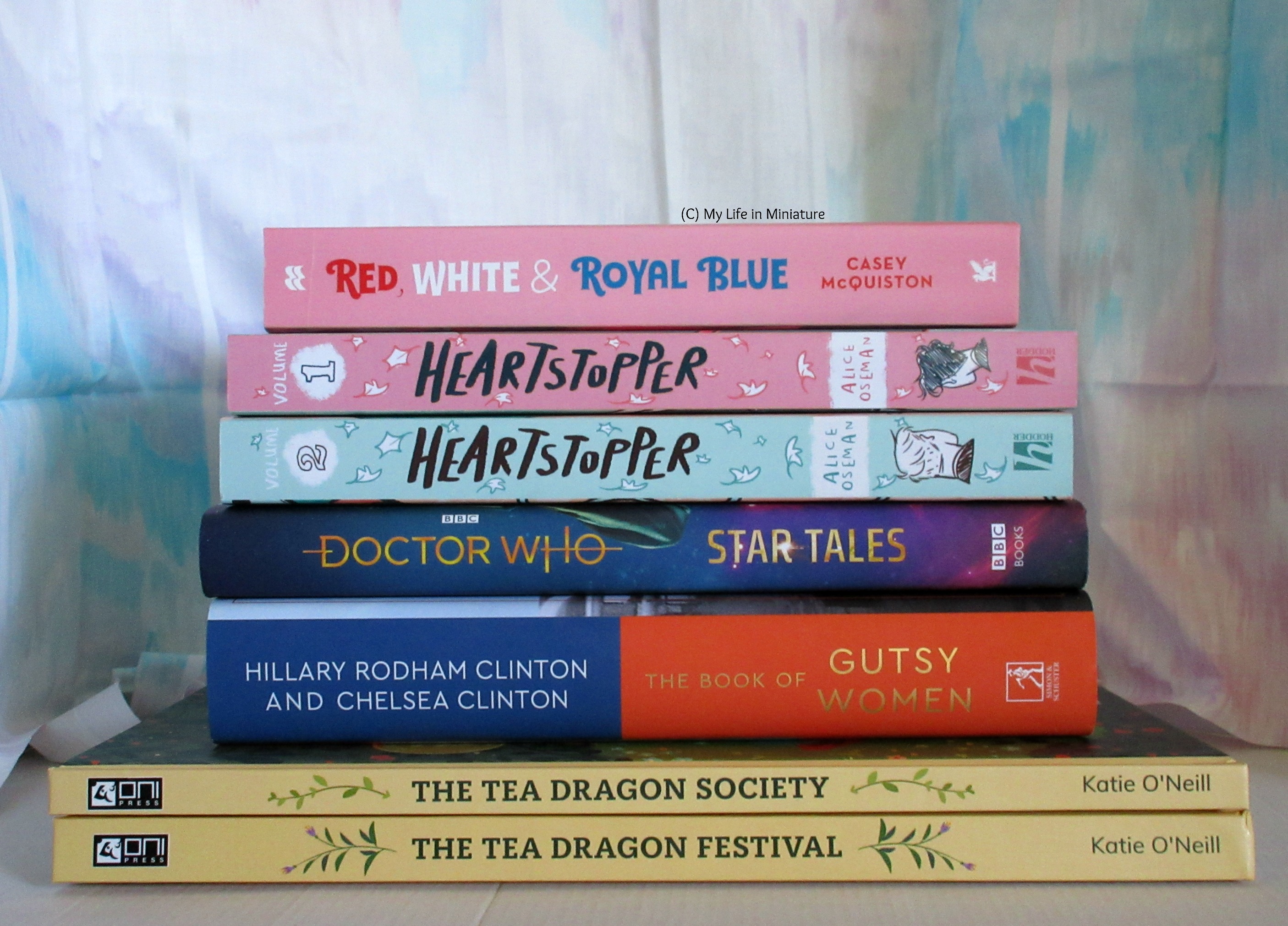 A stack of books, spines facing the camera. Top to bottom, the books are: Red, White and Royal Blue by Casey McQuiston, Heartstopper volumes 1 and 2 by Alice Oseman, Doctor Who Star Tales anthology, The Book of Gutsy Women by Hillary Rodham Clinton and Chelsea Clinton, The Tea Dragon Society and The Tea Dragon Festival by Kay O'Neill.