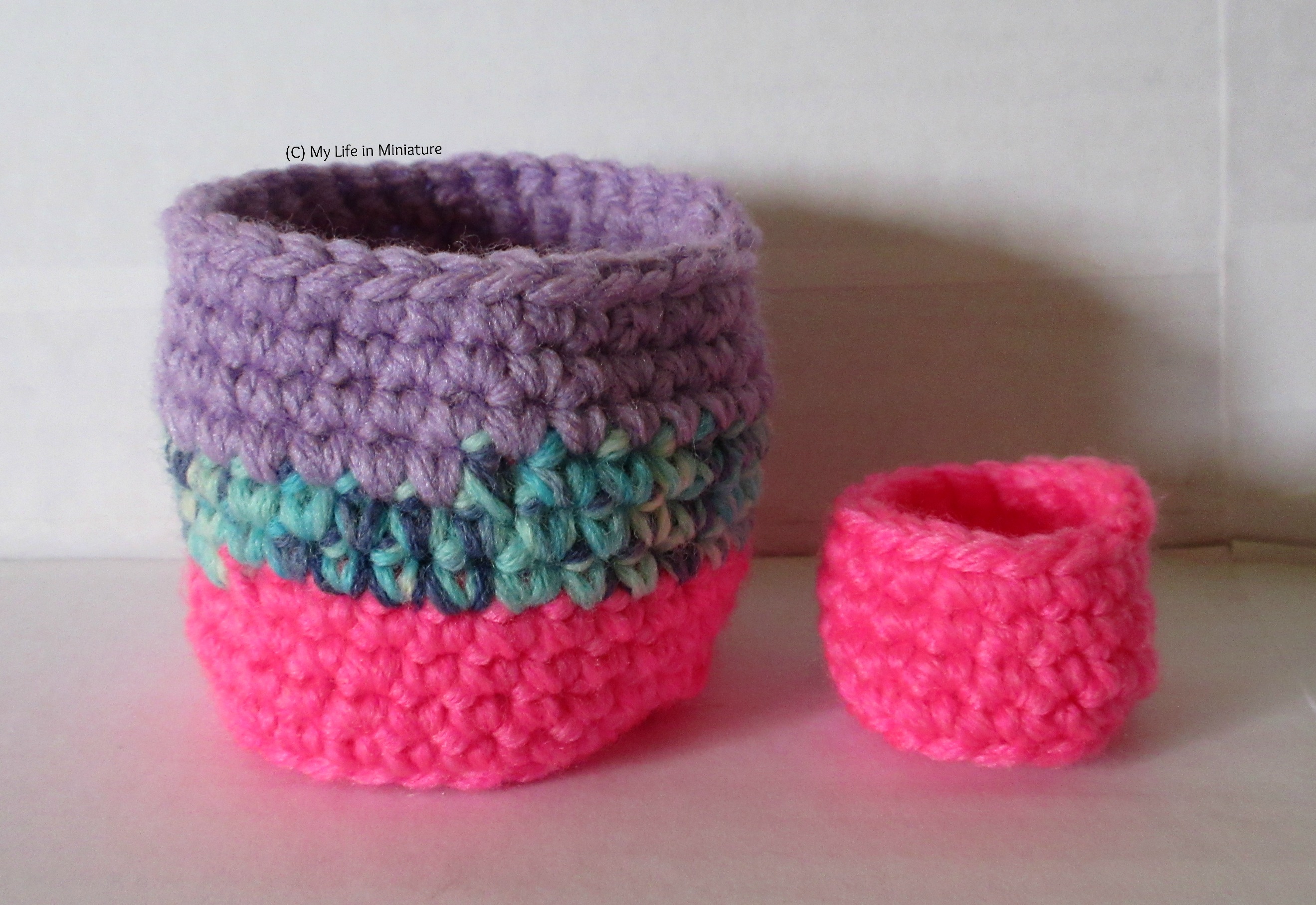 Two crocheted birds' nest/cup-shaped objects stand side-by-side against a white background. One is larger and made of hot pink, variegated blue and plain purple yarn. The other is smaller and hot pink.