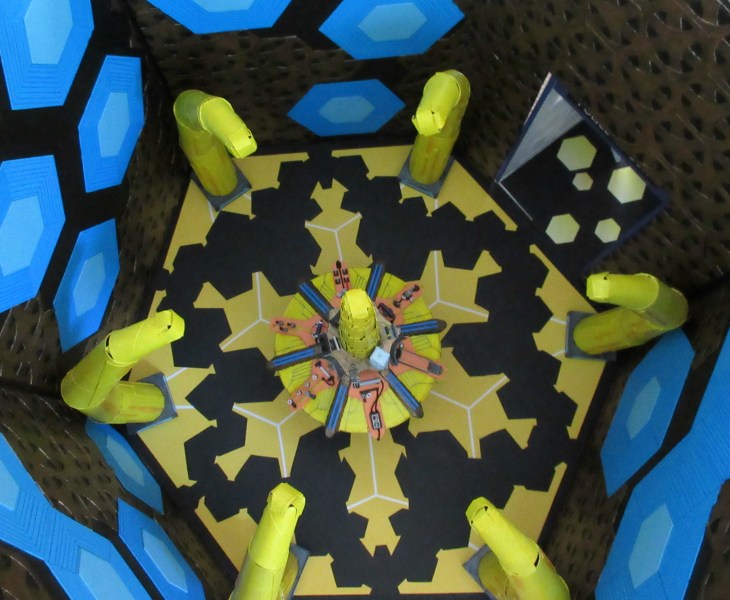 Overhead shot of the interior of the miniature replica of the Thirteenth Doctor's TARDIS. The floor is hexagonal, with yellow and black designs on it, and six yellow arching pillars around the edge. The console rises in the middle, again with six sides full of gadgets and gizmos. The walls are covered in small bronze hexagons and larger blue hexagons. The TARDIS foyer is to the top right.