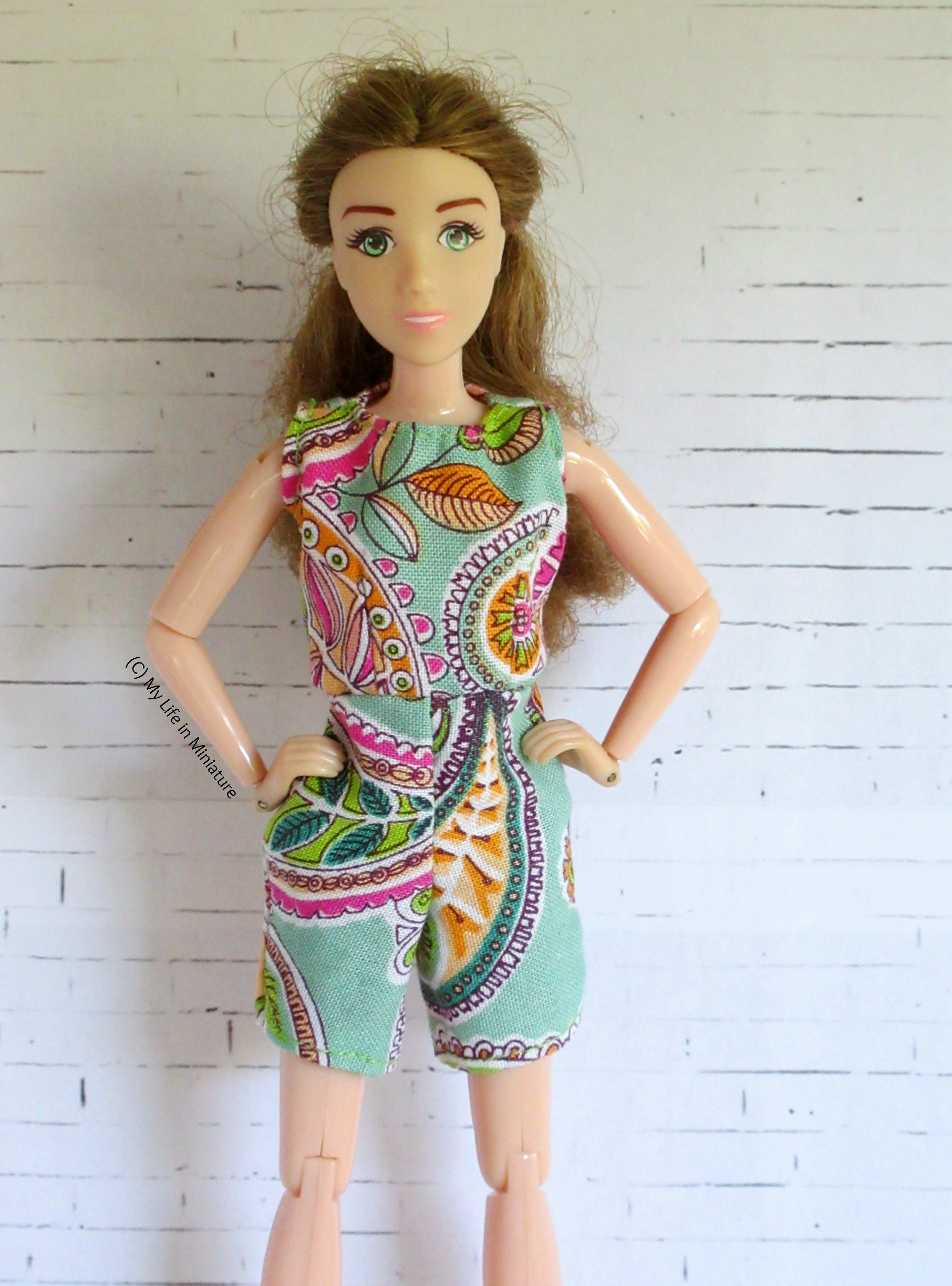 Sarah wears her new playsuit against a white brick background, hands on hips. The fabric is pale green with pink, green and orange paisleys on. 