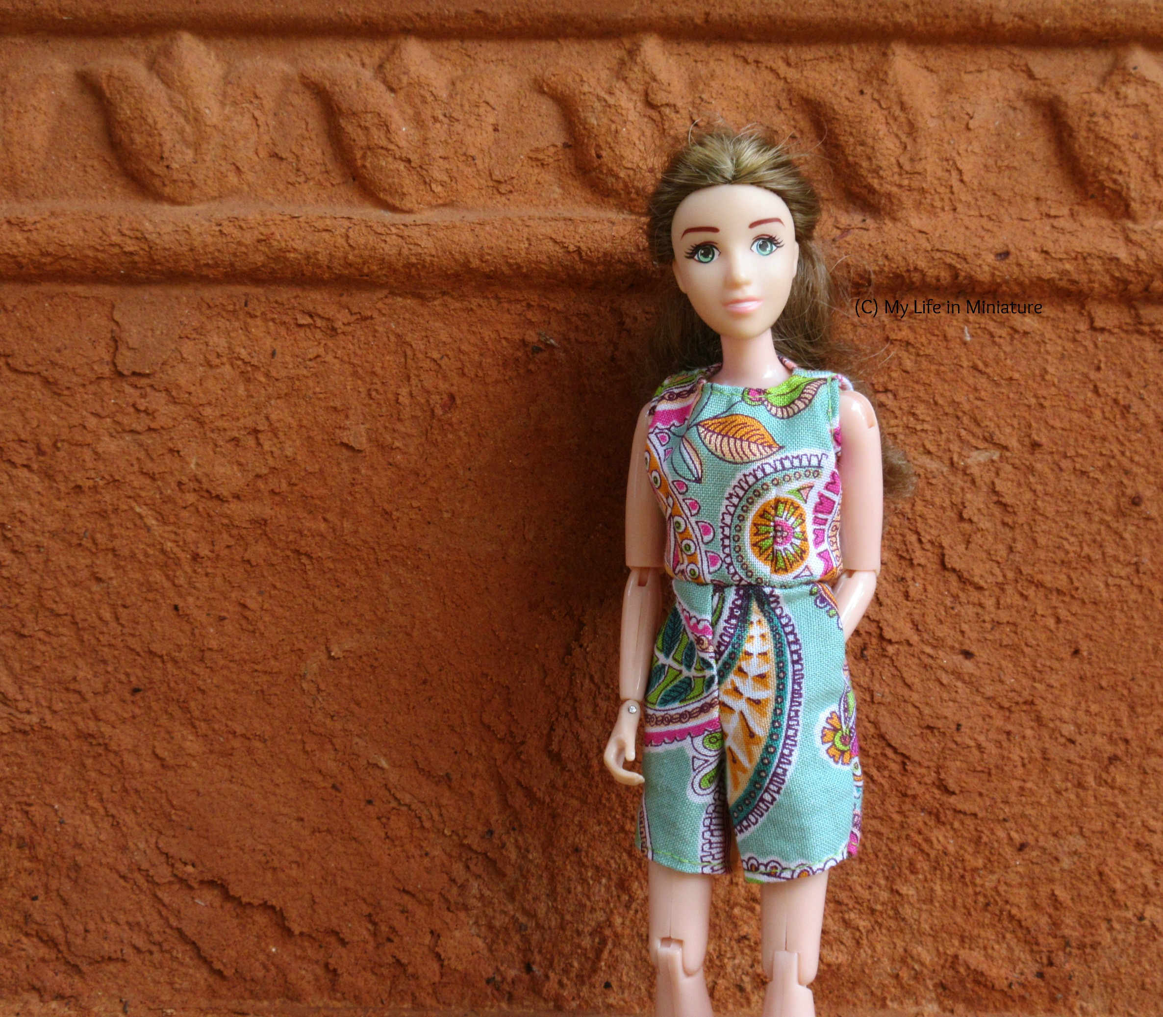 Sarah wears the playsuit with one hand in her pocket and the other hanging beside her. She looks at the camera, and is standing against a rough terracotta background. 