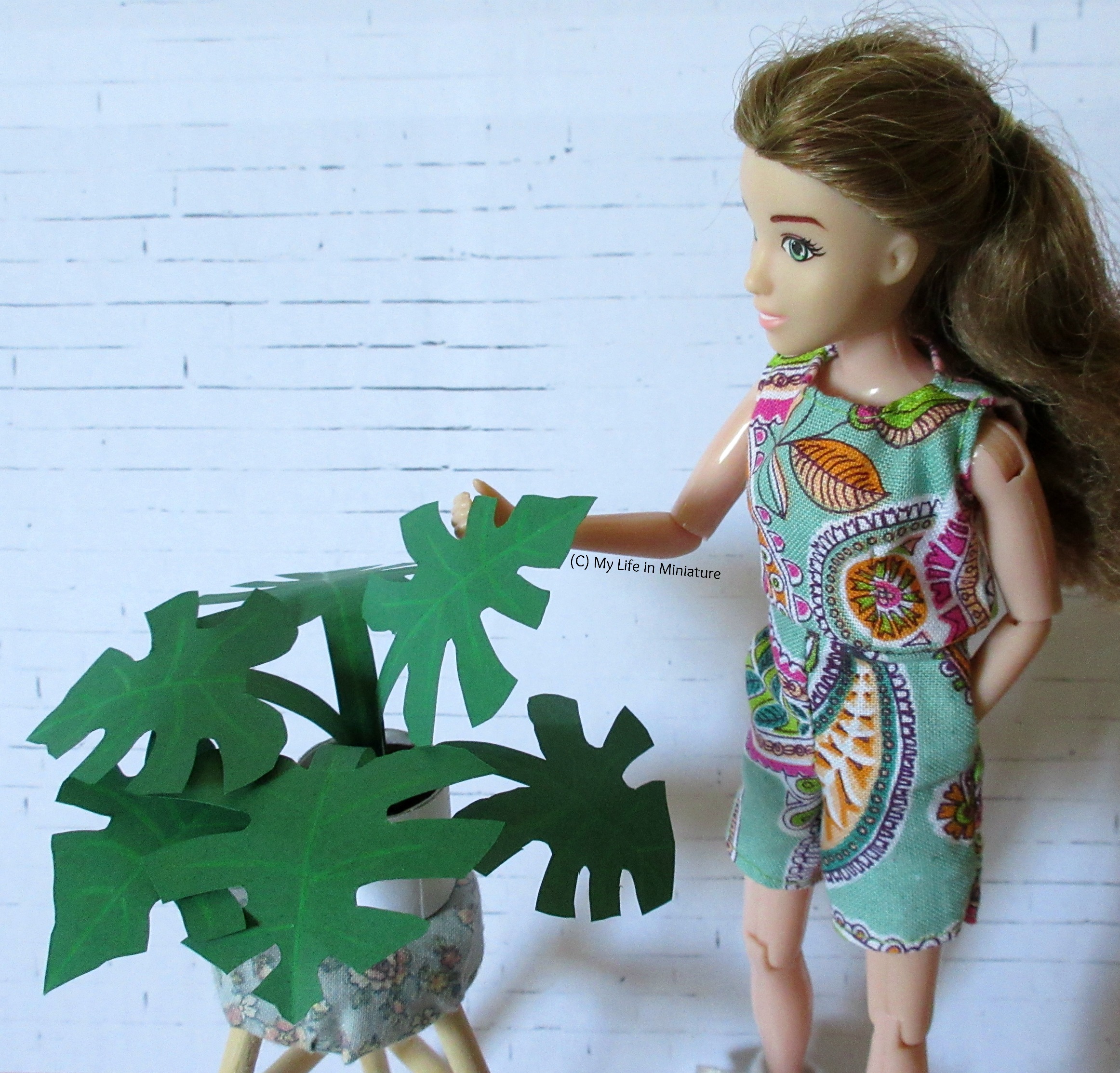 The monstera plant sits on a stool next to Sarah. who is examining one of its leaves. 