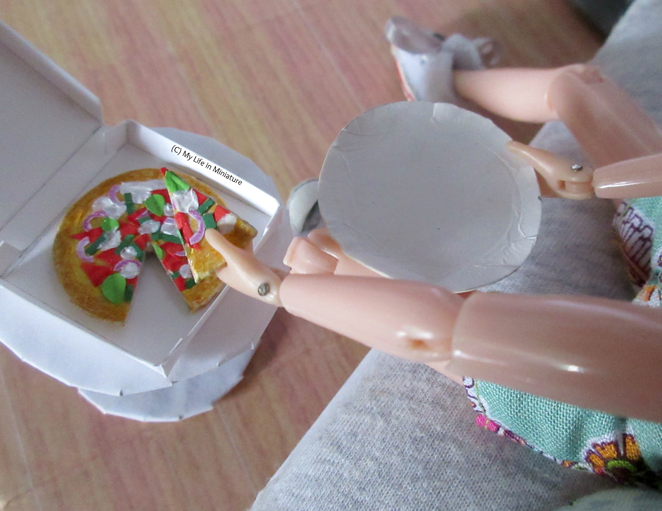 Sarah sits on her couch, one hand holding a plate and the other lifting a slice of vegetarian pizza from the box. The box sits on a small white side table near the couch. The image is taken from above. 
