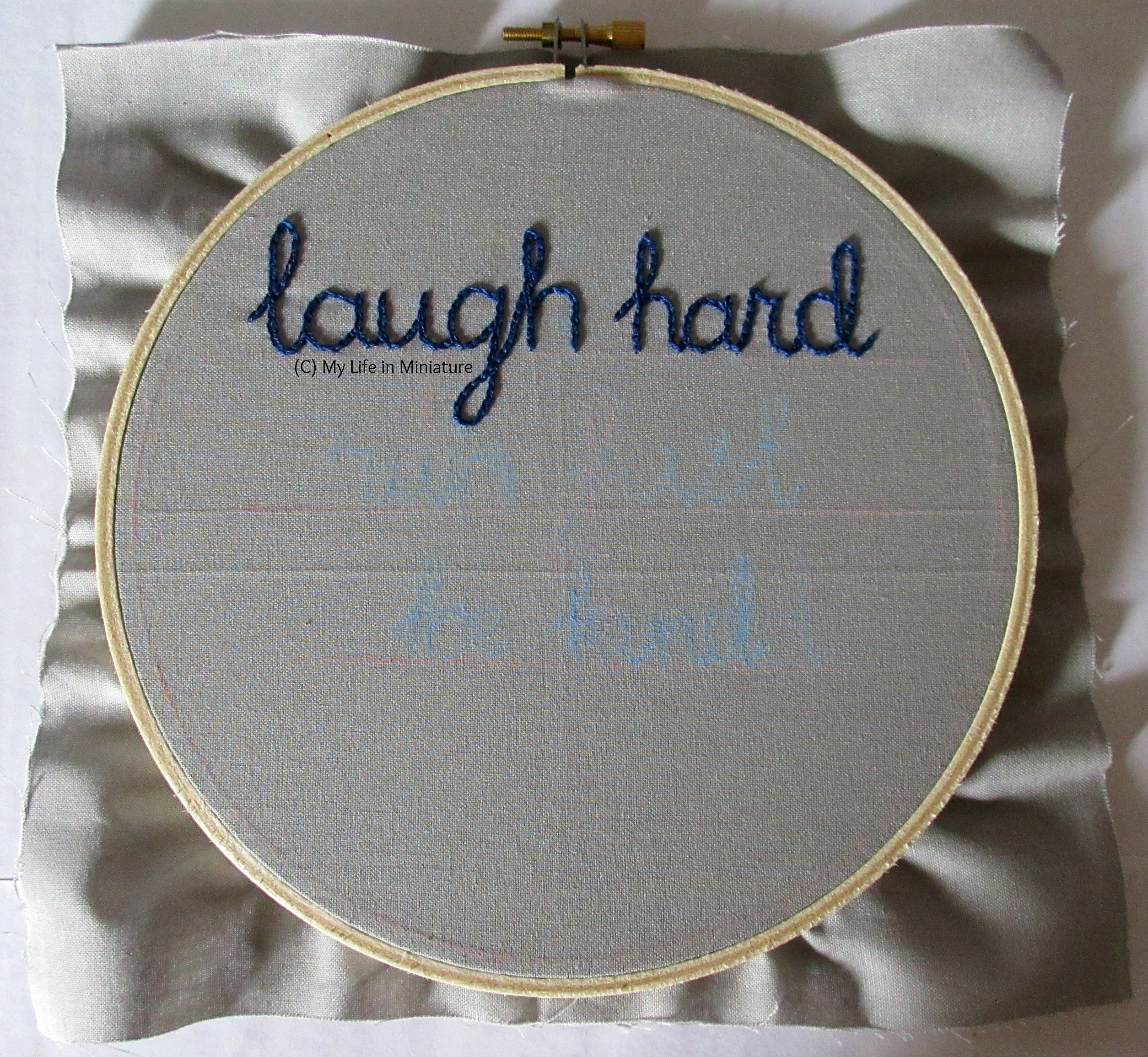 An embroidery hoop fills the image, strung with grey fabric. The phrase 'laugh hard' is stitched across the top third in dark blue chain stitch.