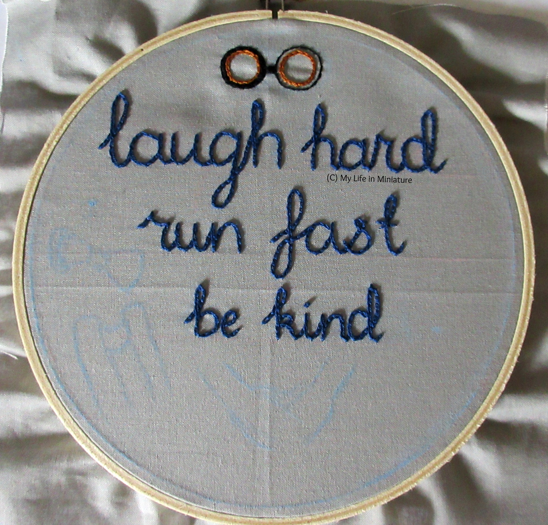An embroidery hoop fills the image, strung with grey fabric. The phrase 'laugh hard; run fast; be kind' is stitched across the top two-thirds in dark blue chain stitch. Above 'laugh hard' is the beginnings of the eyepieces of some steampunk black and gold goggles. 