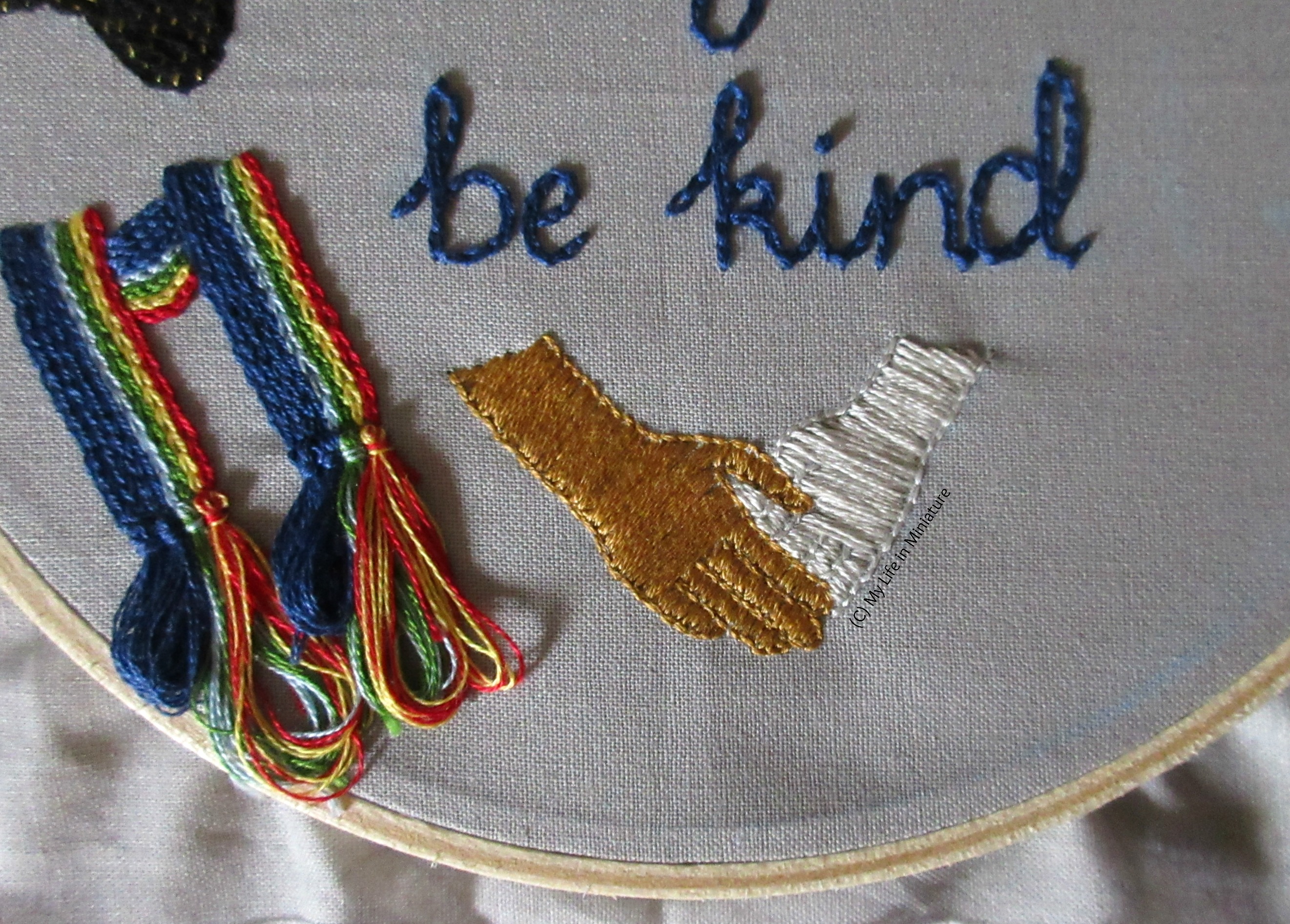 Shot from further back of the bottom of the grey hoop. The scarf tassels are visible on the left, and the two hands are now both filled in. The phrase 'be kind' is visible in dark blue above the hands.