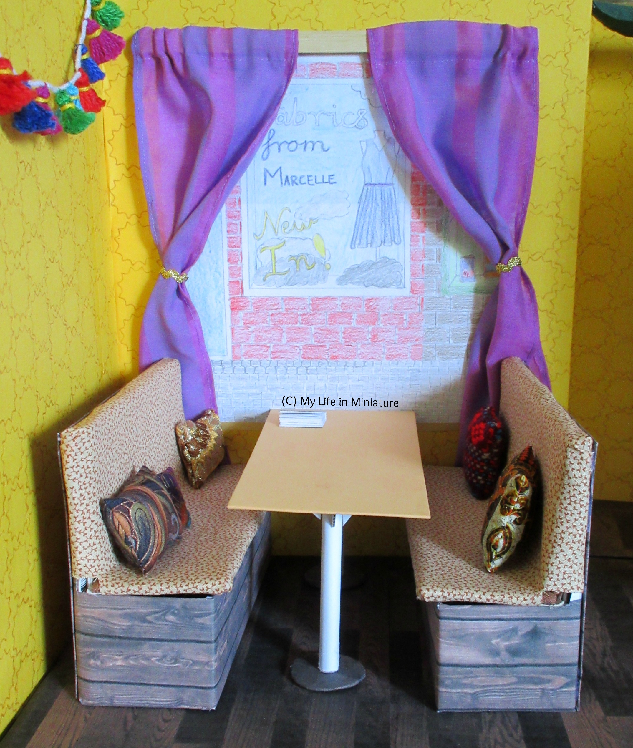 The booth area of Tea at Yaz's is visible. There are two purple curtains over the front window, pulled aside with metallic gold bric-a-brac. The curtains are on a wooden rail.