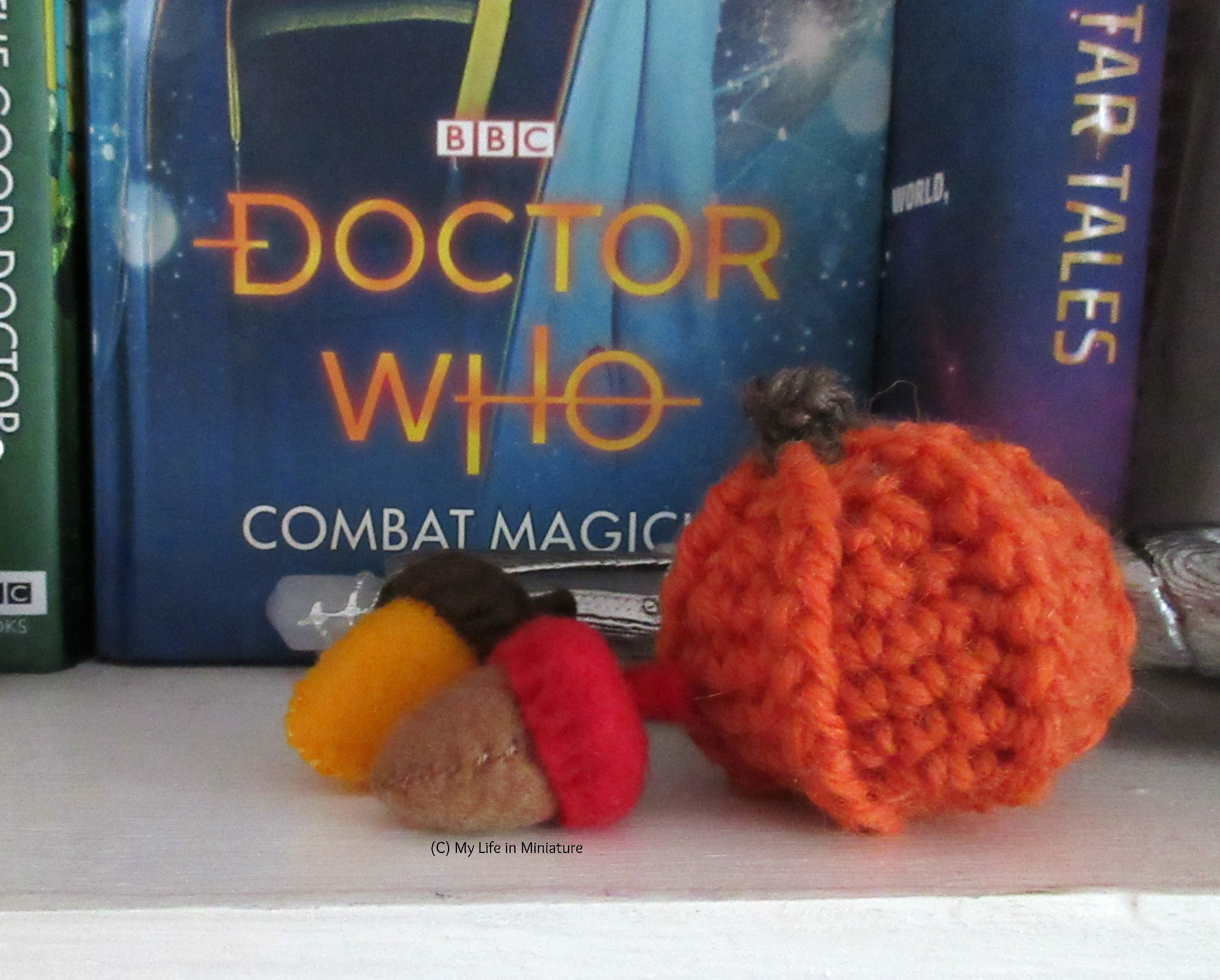 Two felt acorns sit on a bookshelf, next to a crocheted orange pumpkin. 