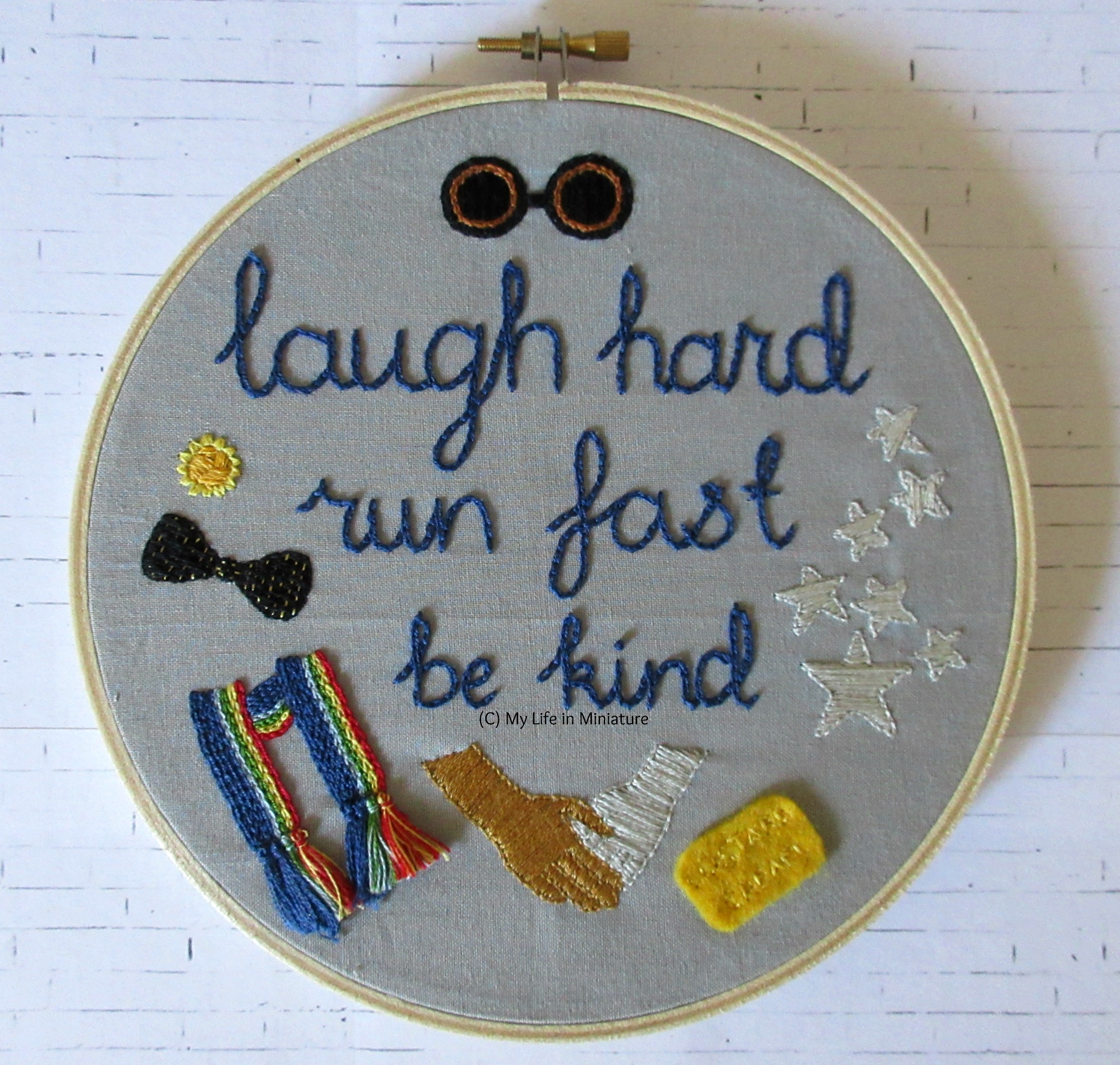 The embroidery hoop, filled with grey fabric, sits on a white brick background. The phrase 'laugh hard; run fast; be kind' is embroidered in navy blue in the middle. Around it are various symbols of the Thirteenth Doctor, including a daffodil flower, a rainbow-striped scarf, and a custard cream biscuit. 