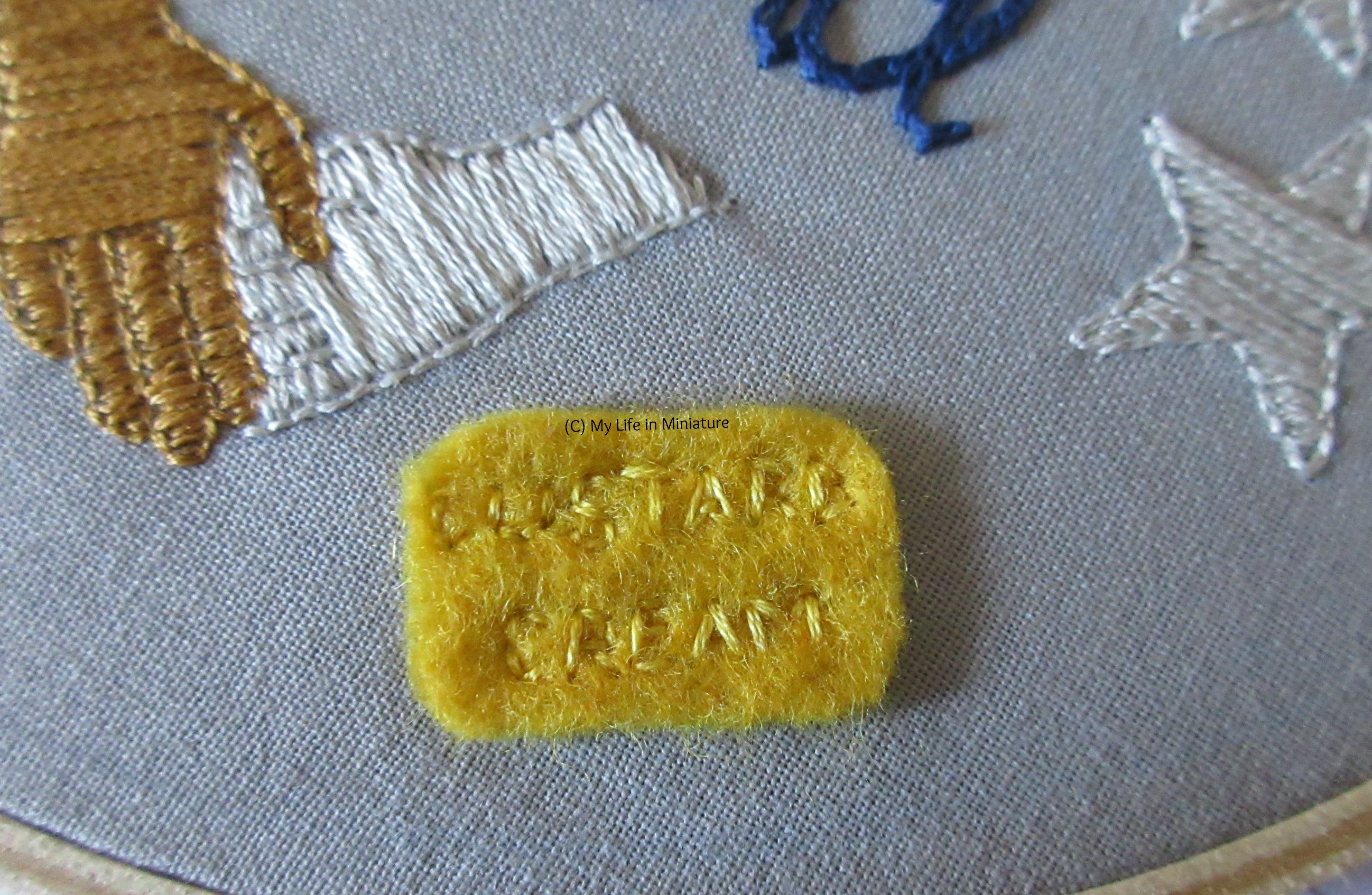 Close-up of a rectangle of yellow felt on the hoop. Stitched into it in yellow are the words 'custard cream', to look like the biscuit. 