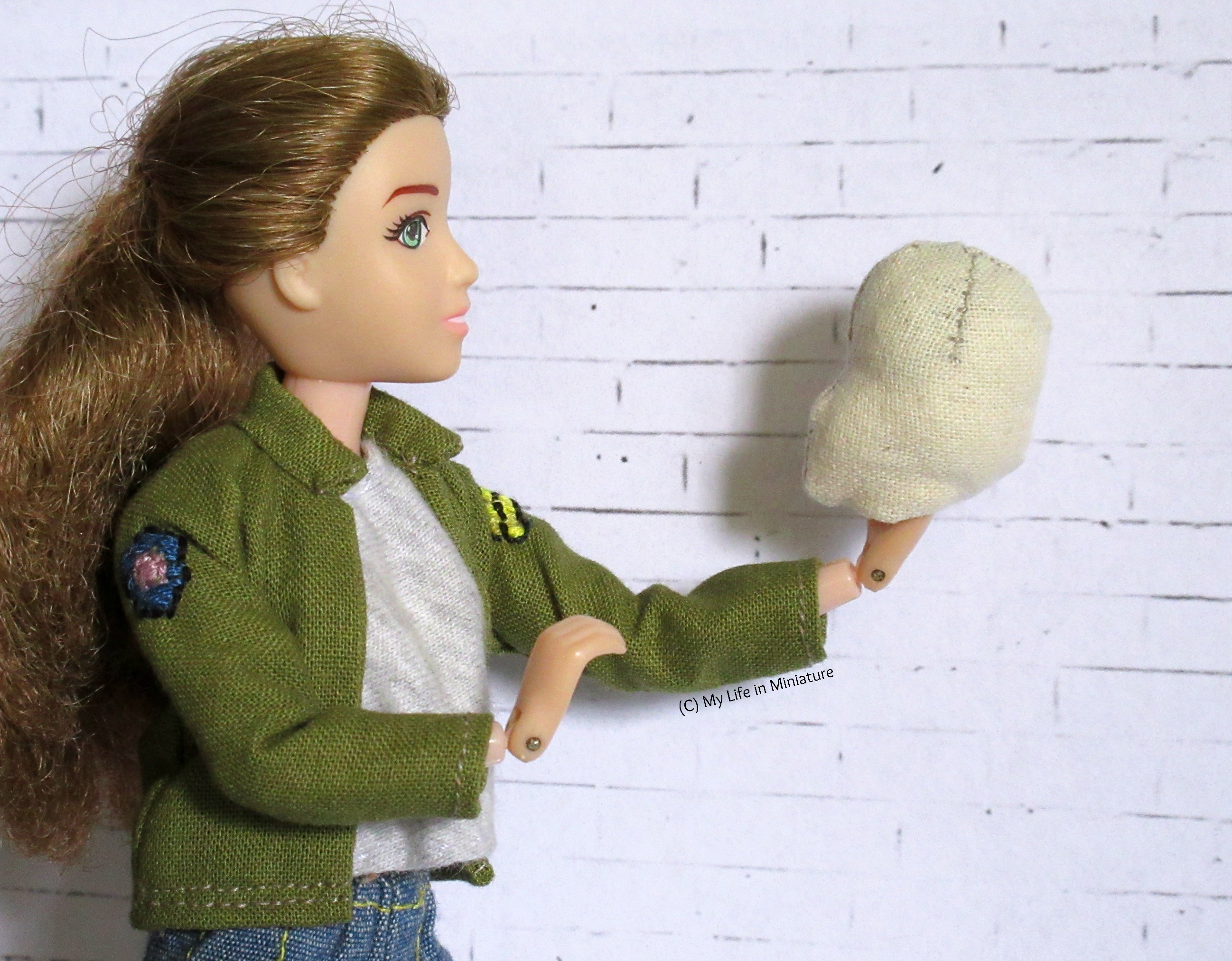 Sarah holds the stuffed head of the muslin doll in one hand. She holds it out away from herself, with the head looking back at her. She looks at the head. The whole pose is reminiscent of Hamlet. 