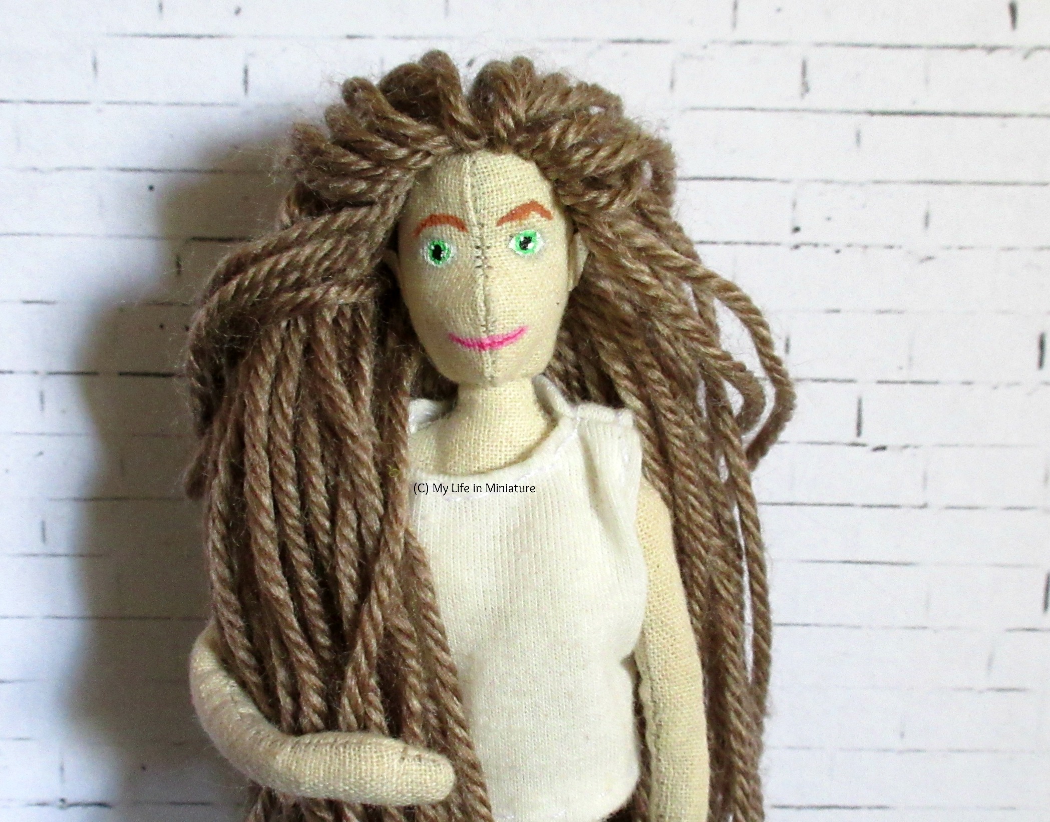 A handmade muslin doll stands in front of a white brick background. She wears a white t-shirt, and has pale brown hair made of yarn and green eyes.