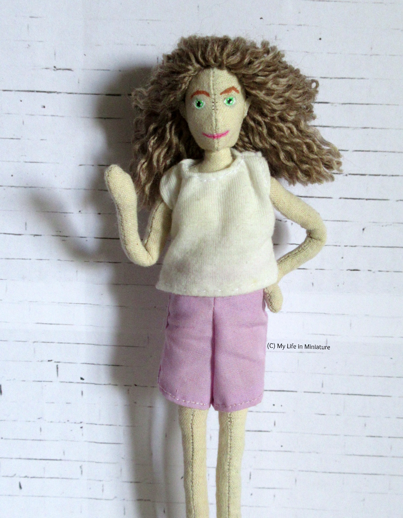 The doll waves at the camera, looking at it. Her hair is shorter, about shoulder-blade length. She wears purple shorts. 