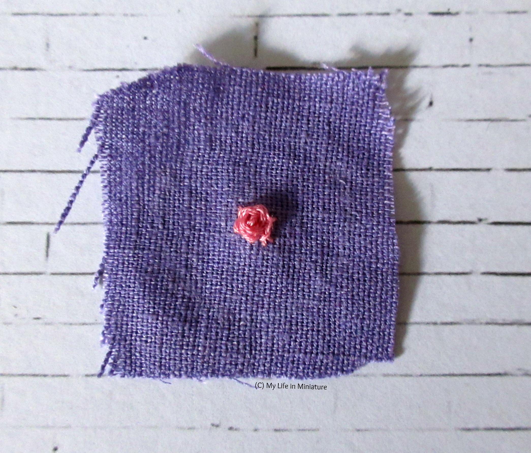 A purple square of fabric sits on a white brick background. A pink rose is embroidered in the centre. 