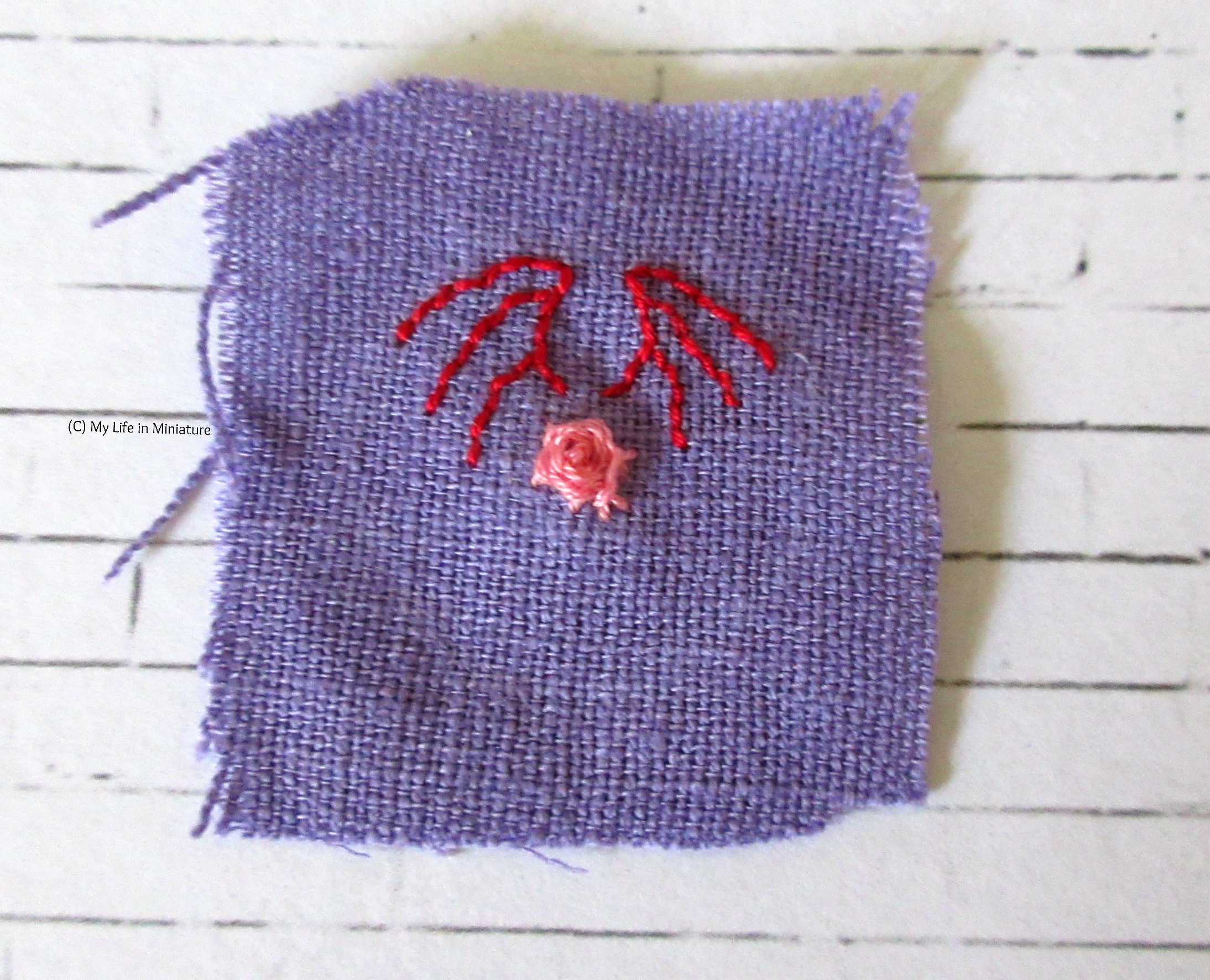 A square of purple fabric sits on a white brick background. Above the pink rose in the centre is embroidered the outline of two red dragon wings. 