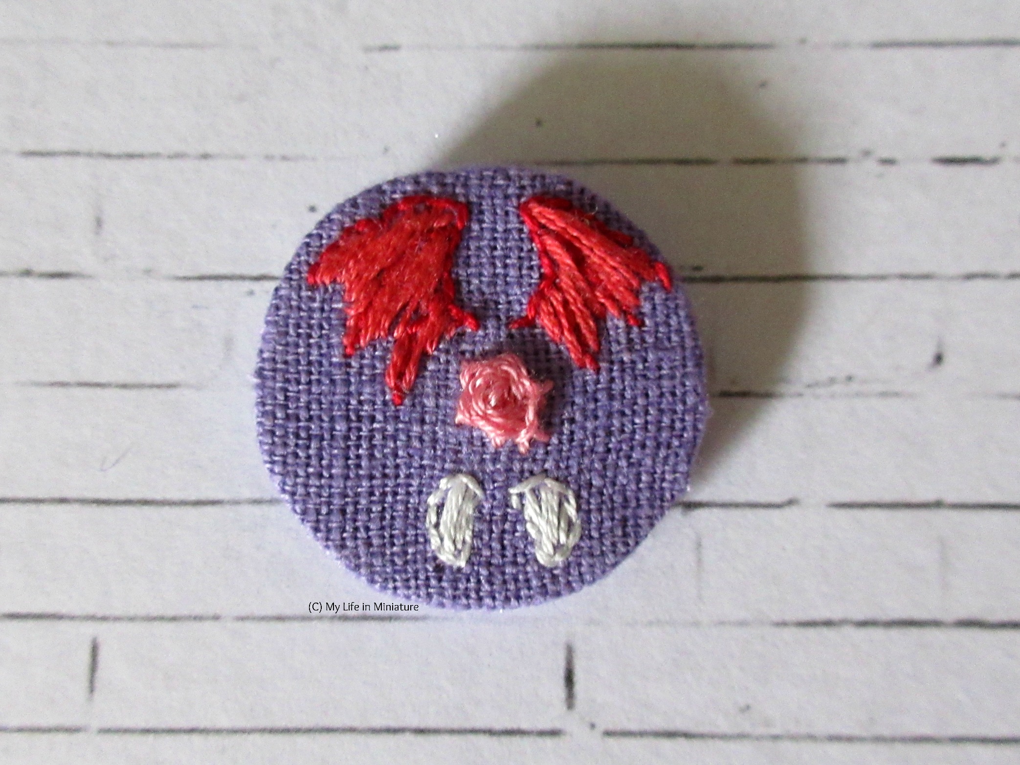 The finished circular badge sits on a white brick background. A pink rose is embroidered in the centre, below which are two vampire teeth. Above is a pair of red dragon wings. 