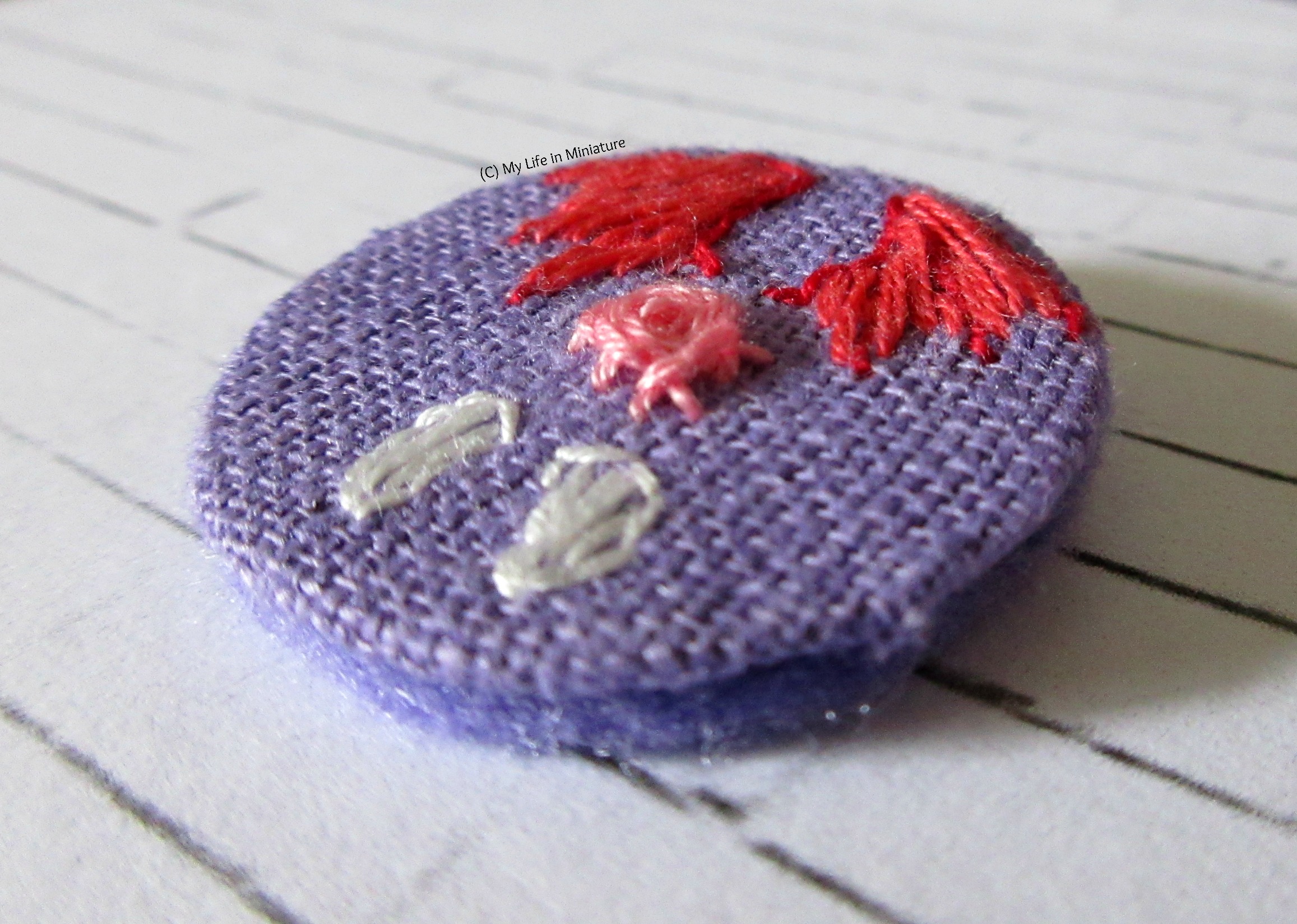 Close-up shot of the badge from an angle to show the elevation/texture of the stitches. It sits on a white brick background. 