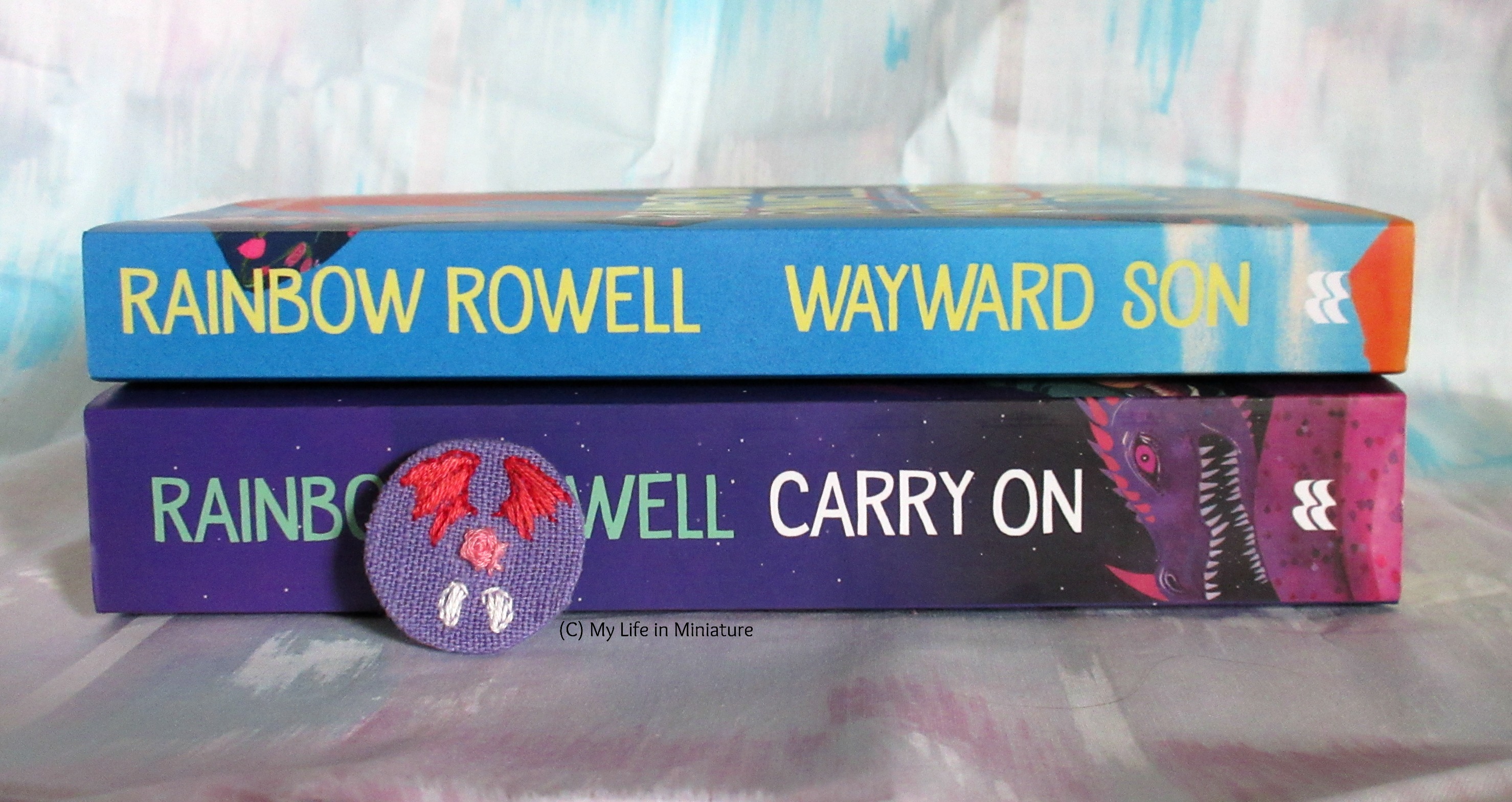 'Carry On' and 'Wayward Son' by Rainbow Rowell sit stacked on each other, spines facing the camera. The badge leans up against the spine of 'Carry On'. 