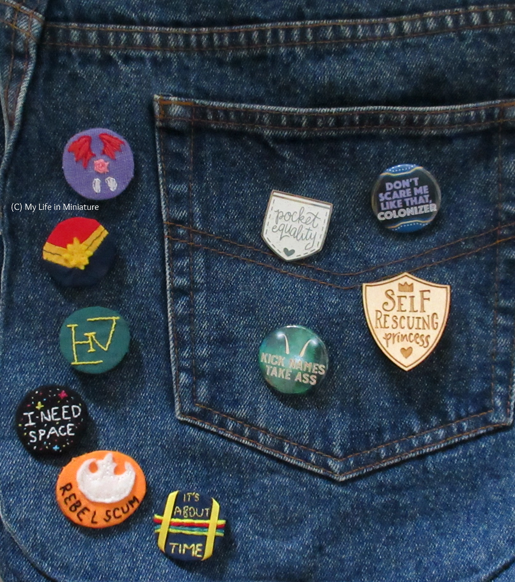 The badge is pinned onto a denim bag, alongside a variety of other badges and pins - both handmade and purchased. 