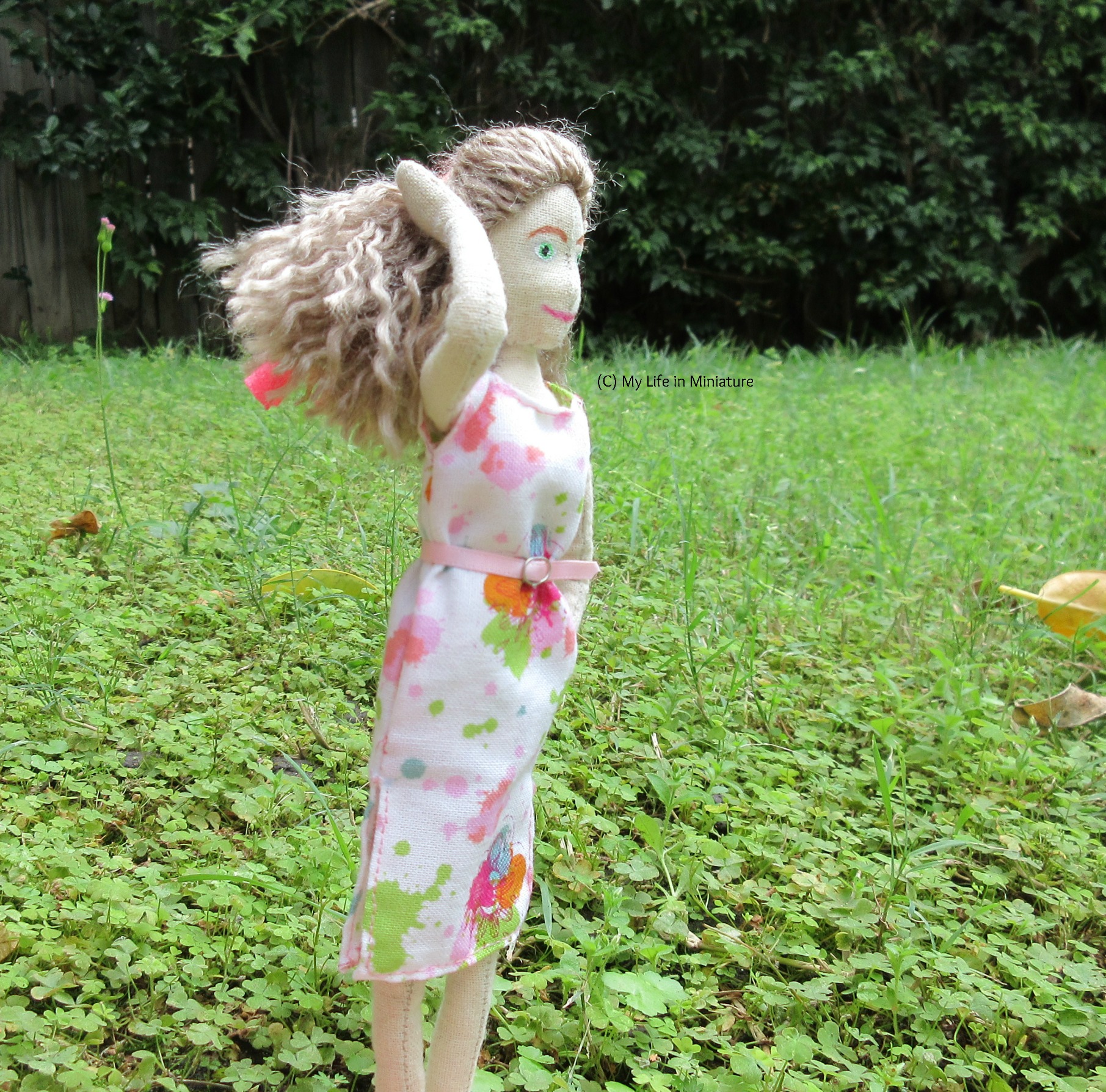 Lola stands on grass, with a hedge behind her. She has one hand on her hair and the other in her pocket, and she looks to the right. 