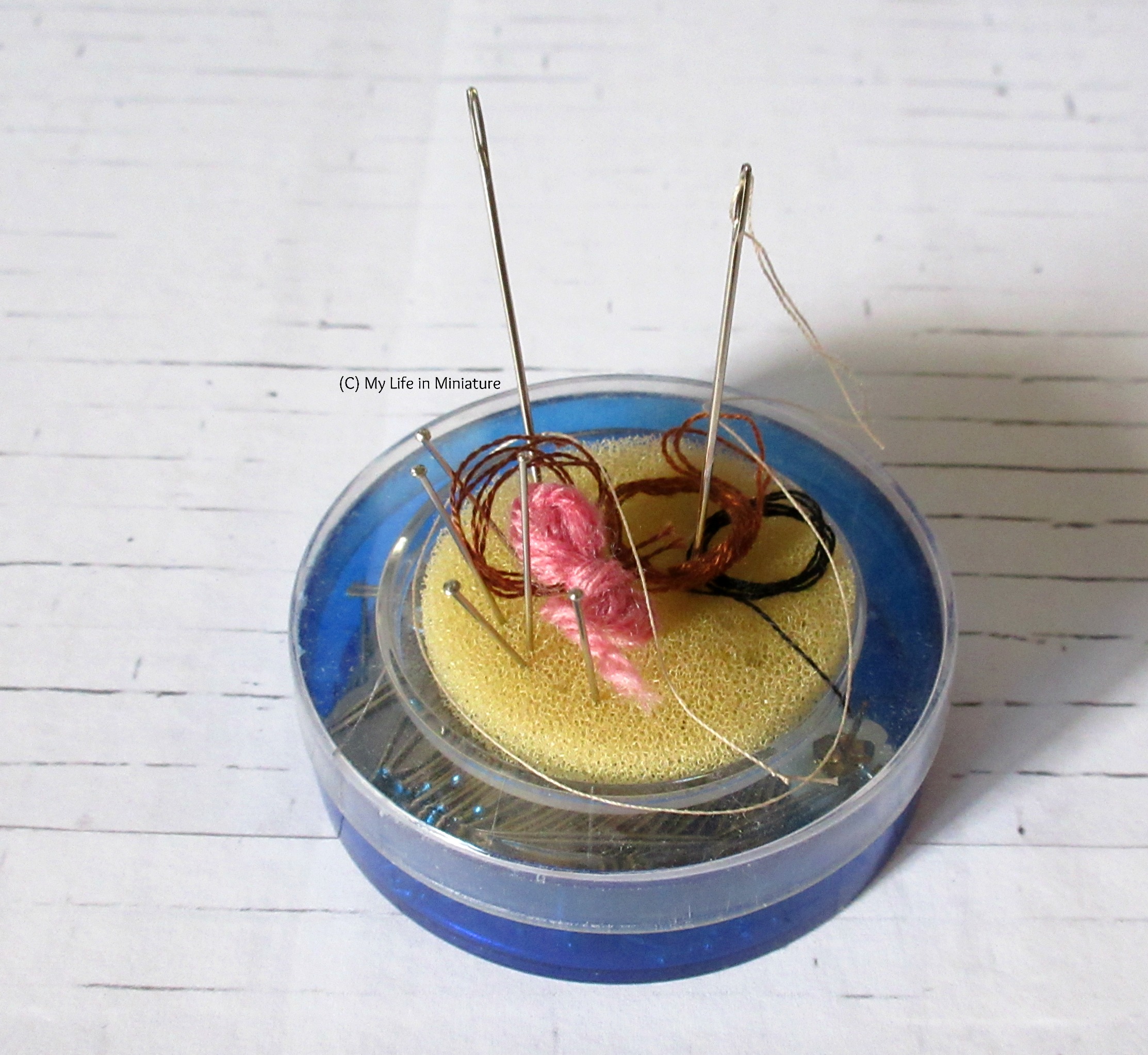 A blue pin tin with a clear plastic lid sits on a white brick background. On top is a yellow circular sponge with needles and pins stuck in it.