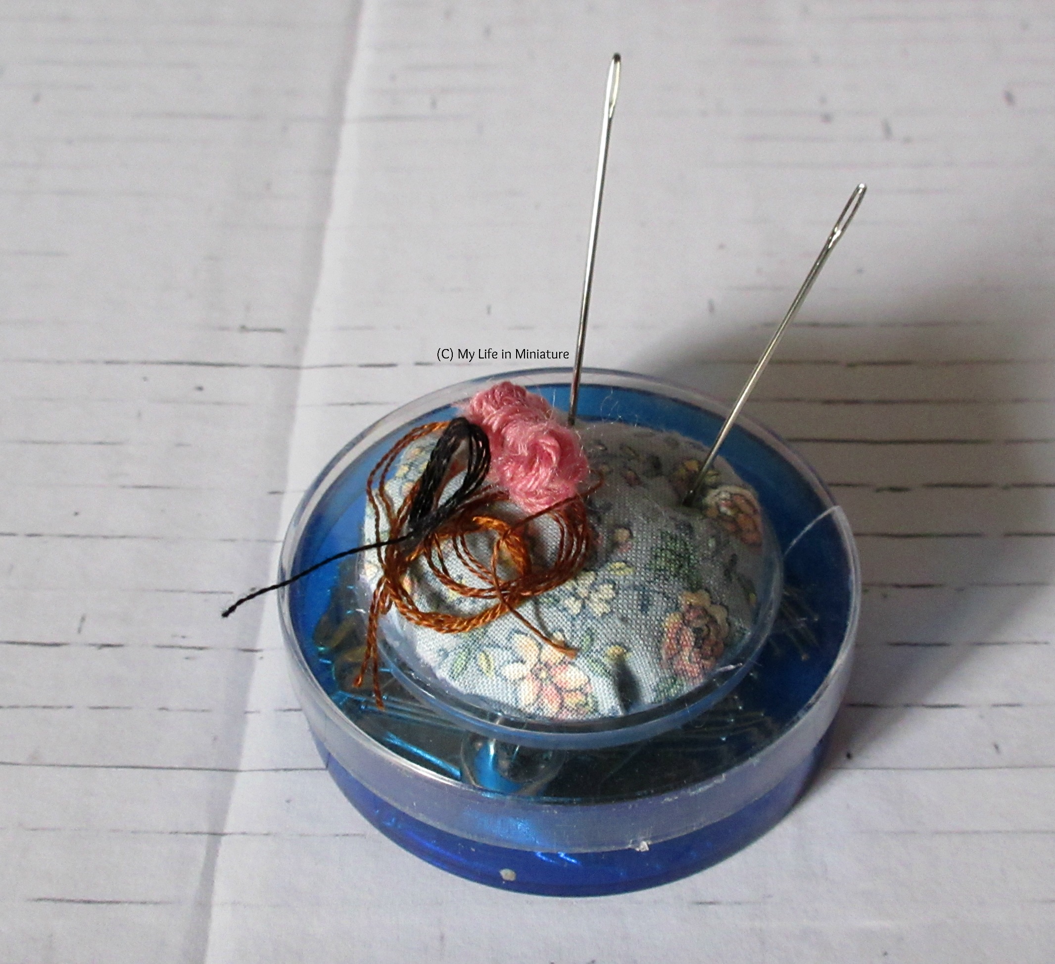 The blue pin tin, with clear lid and blue floral pincushion, sits on a white brick background. There are two needles stuck in it.