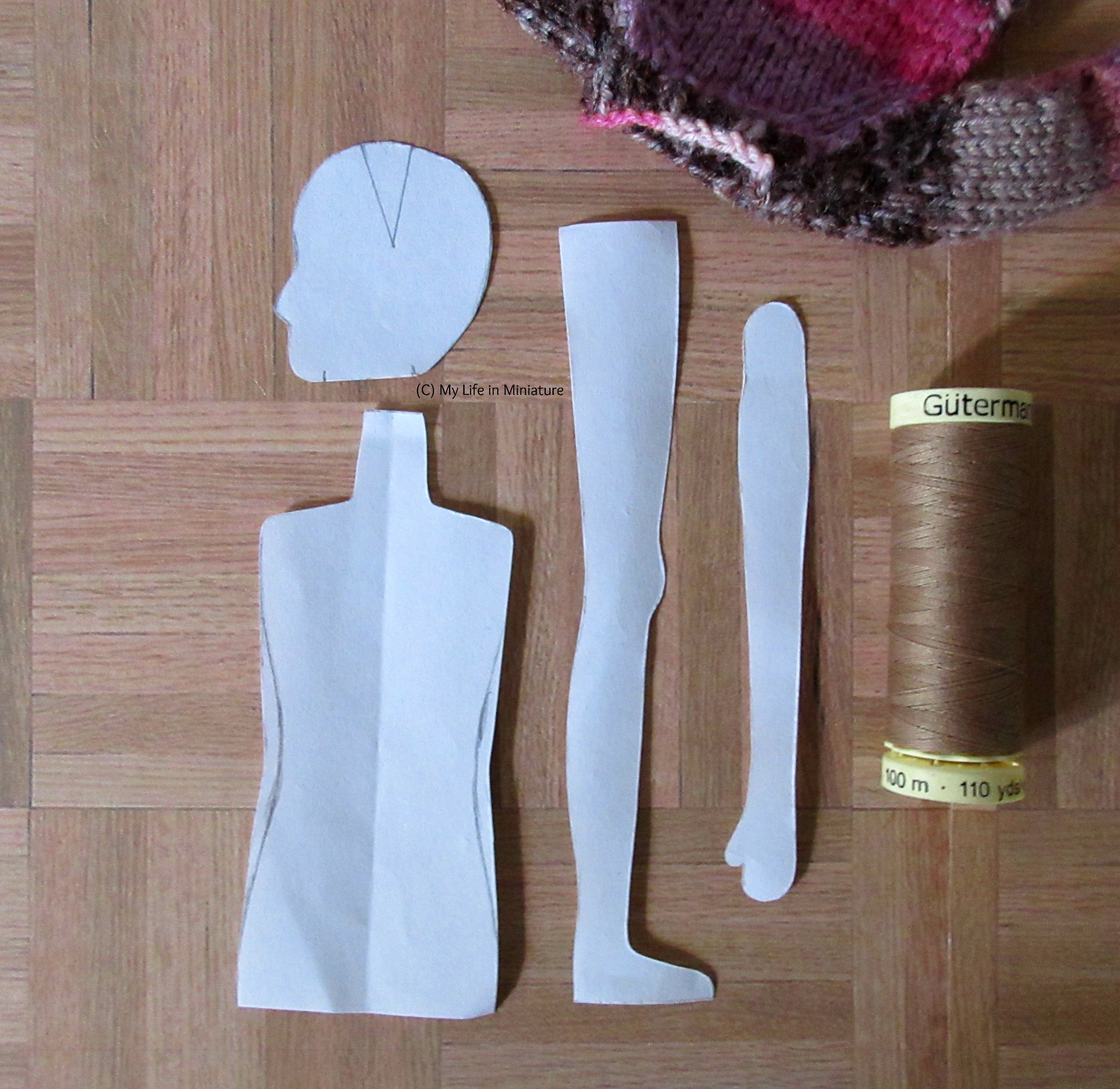 A fabric doll pattern is laid out on a wood parquet background. A reel of thread in pale brown and a knitted pink jumper are next to the pattern pieces.
