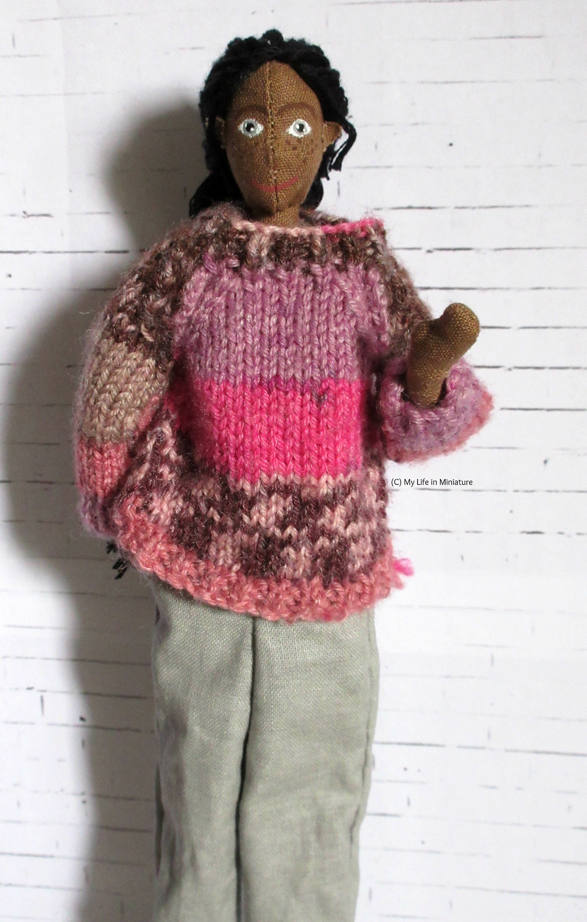 Petra stands against a white brick background. She has brown skin, grey eyes, freckles, and black hair. She wears a pink and purple jumper, loose grey pants, and is waving at the camera. 