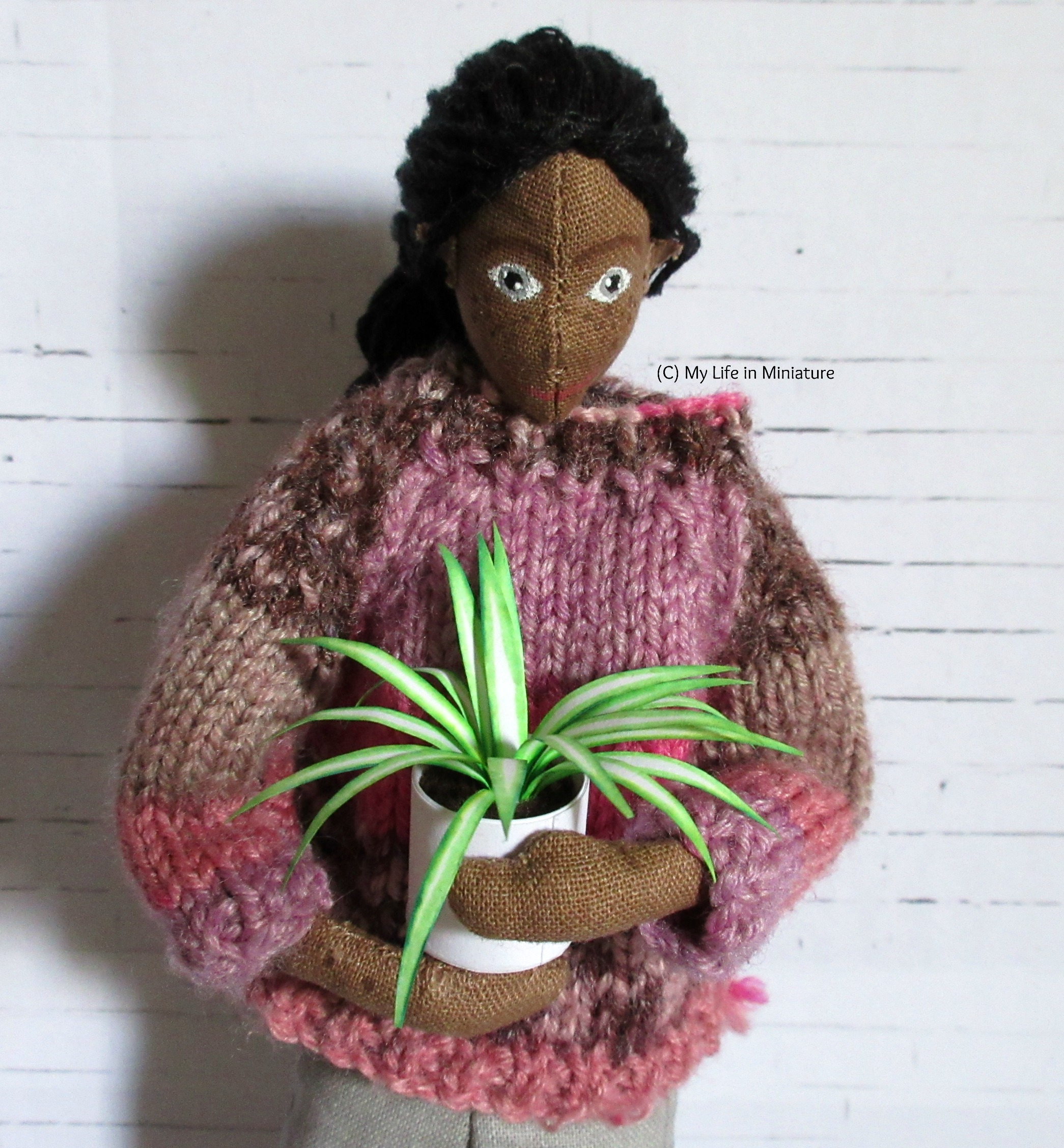 Petra holds a spiderplant in her arms, looking down at it. She's against a white brick background.