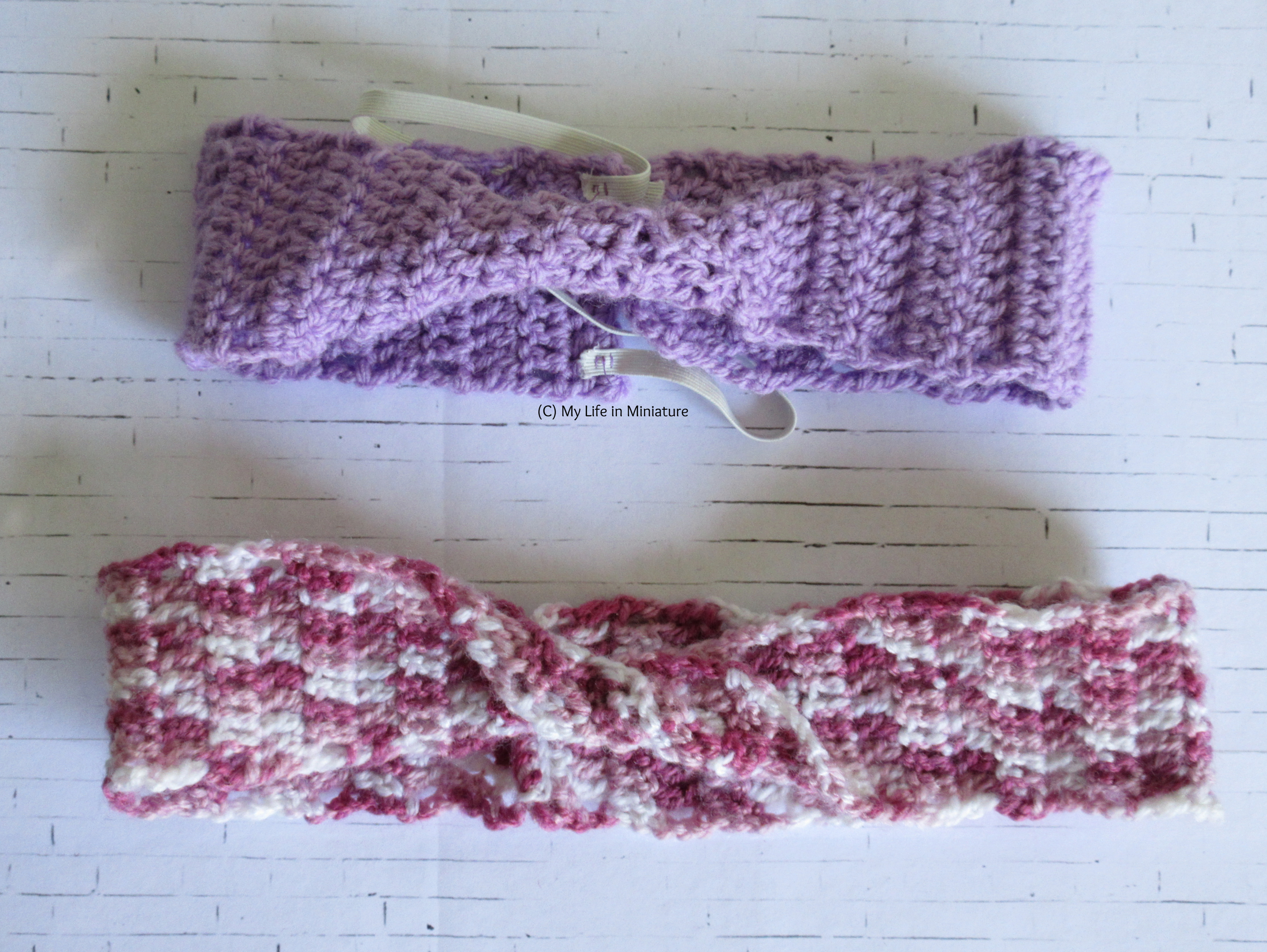 Two crocheted headbands with twists in the centre sit on a white brick background. The top one is pale purple with elastic, and the bottom one is shades of pink and white. 