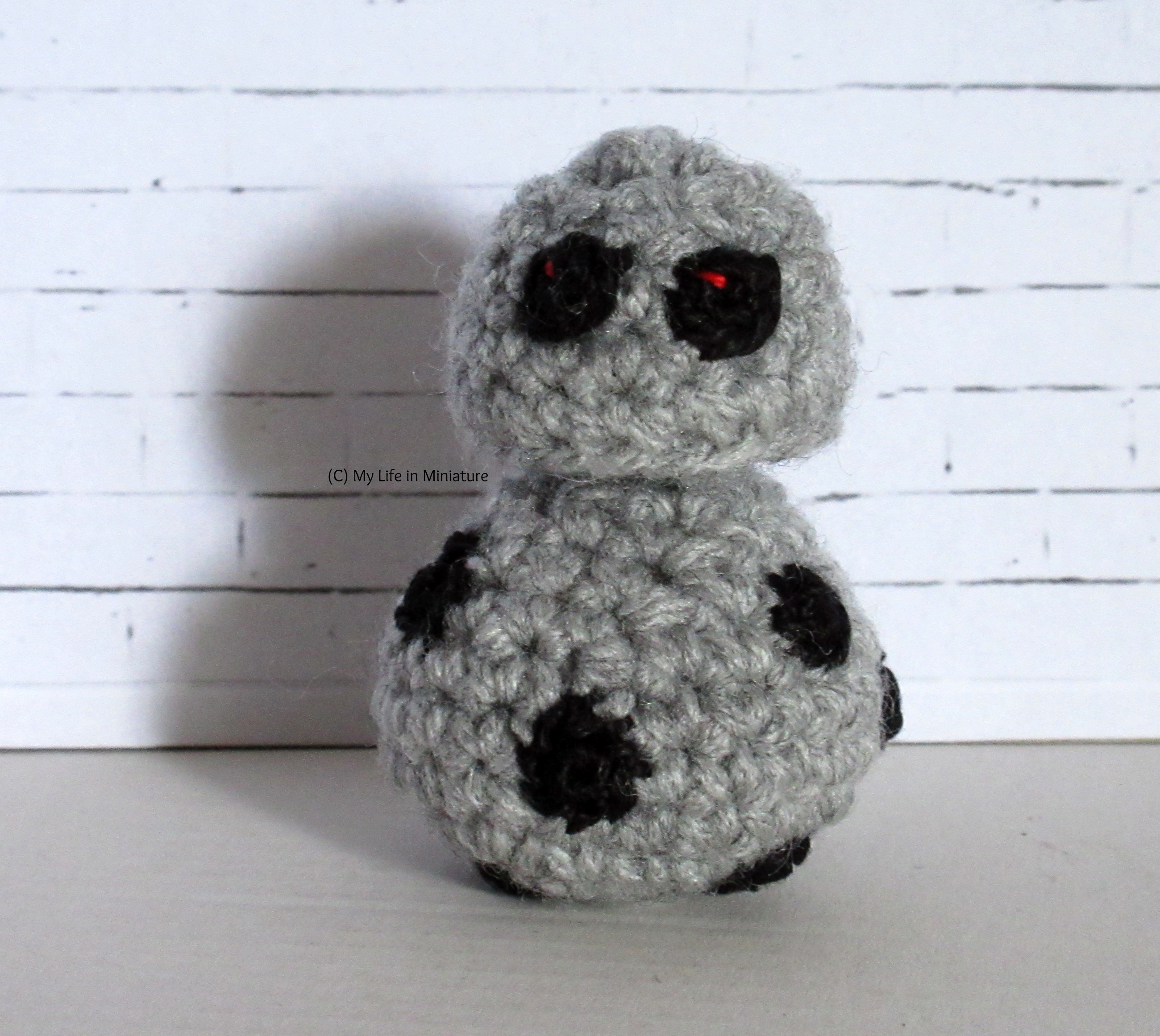 A small android sits against a white brick background. They are the same silhouette as BB-8, but grey with more small black function openings on their body. They have two optical sensors on their head, both looking at the camera.  