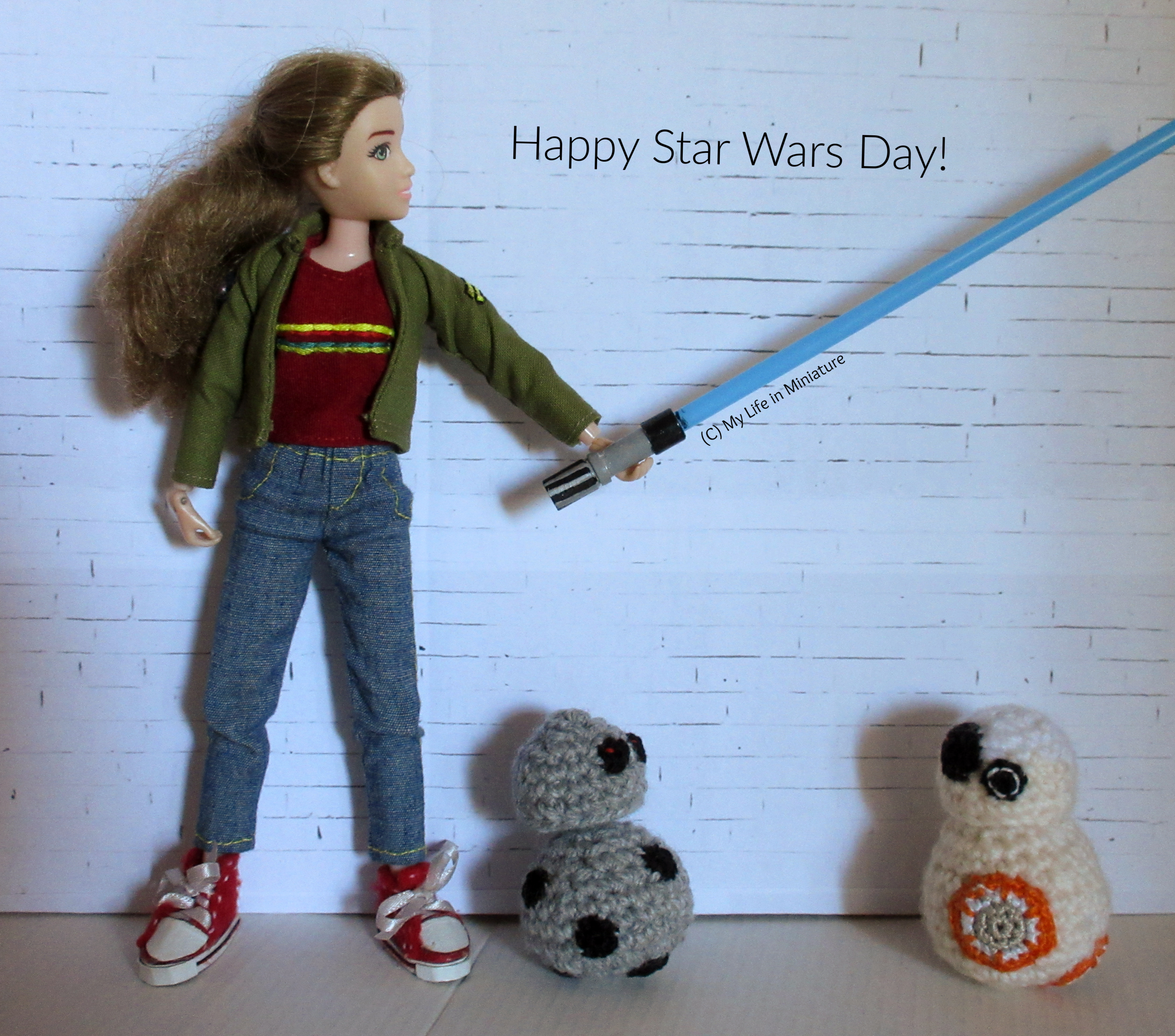 Sarah, BB-8, and Chip stand against a white brick background. Sarah is holding a blue lightsaber out to her side, looking at the laser. Chip and BB-8 are at Sarah's feet, looking at each other. Above the lightsaber are the words 'Happy Star Wars Day!'. 