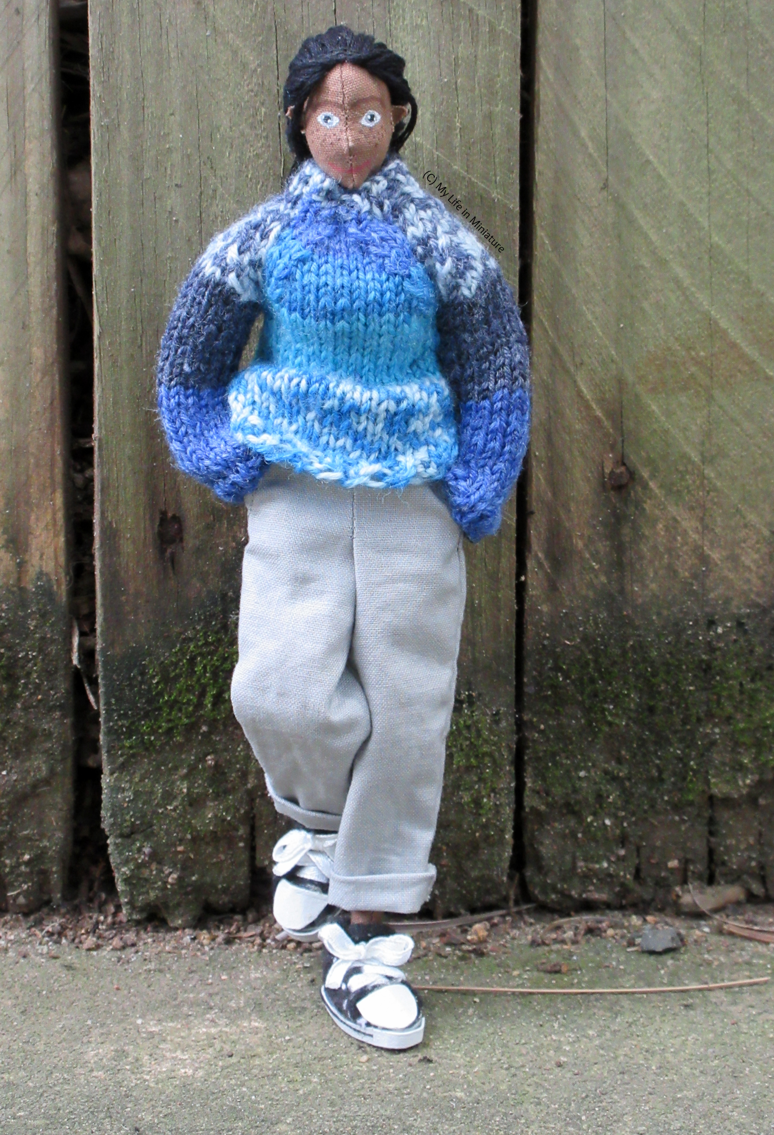 Petra stands against a fence, hands in pockets and foot propped up. She wears the blue jumper and loose grey pants with black Converse high-tops. She's looking at the camera. 