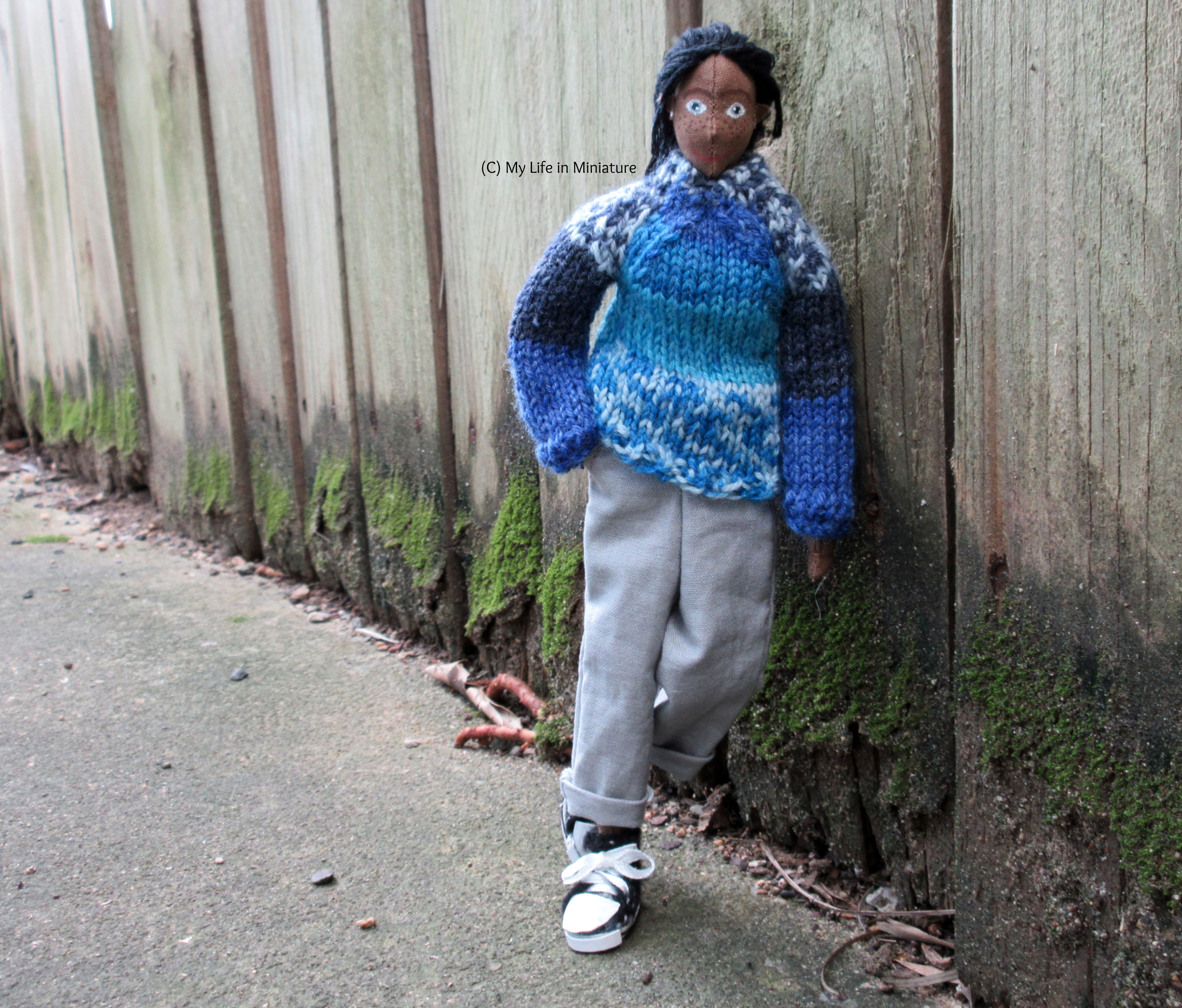 Petra leans a shoulder against a wooden fence, one hand in her pocket and one foot propped up. She's looking at the camera, wearing the blue jumper, grey pants, and black shoes. 