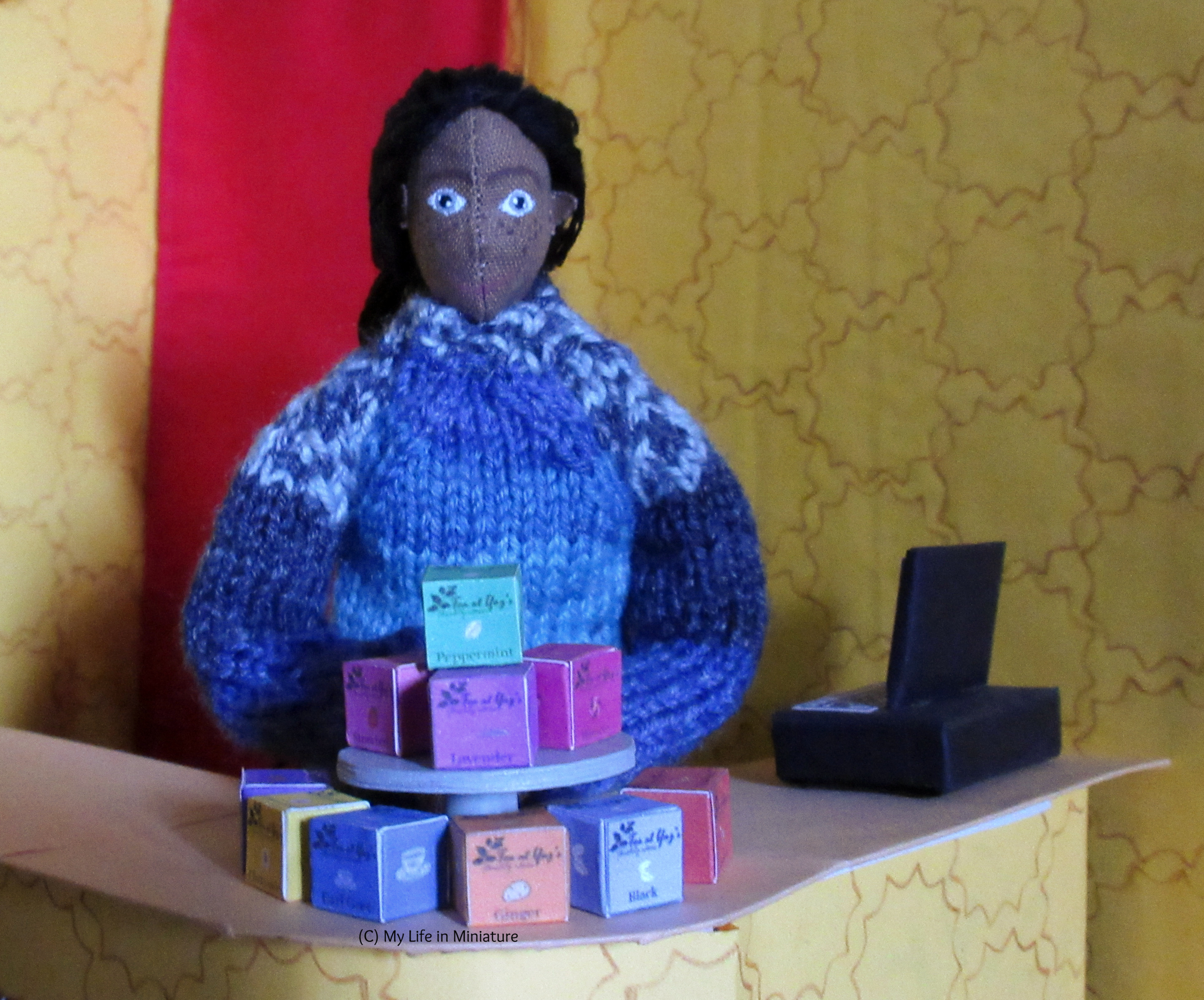 Petra stands behind the cake stand on Tea at Yaz's counter. The cake stand is stacked with tea boxes. She looks at the camera, hands neatly folded. 