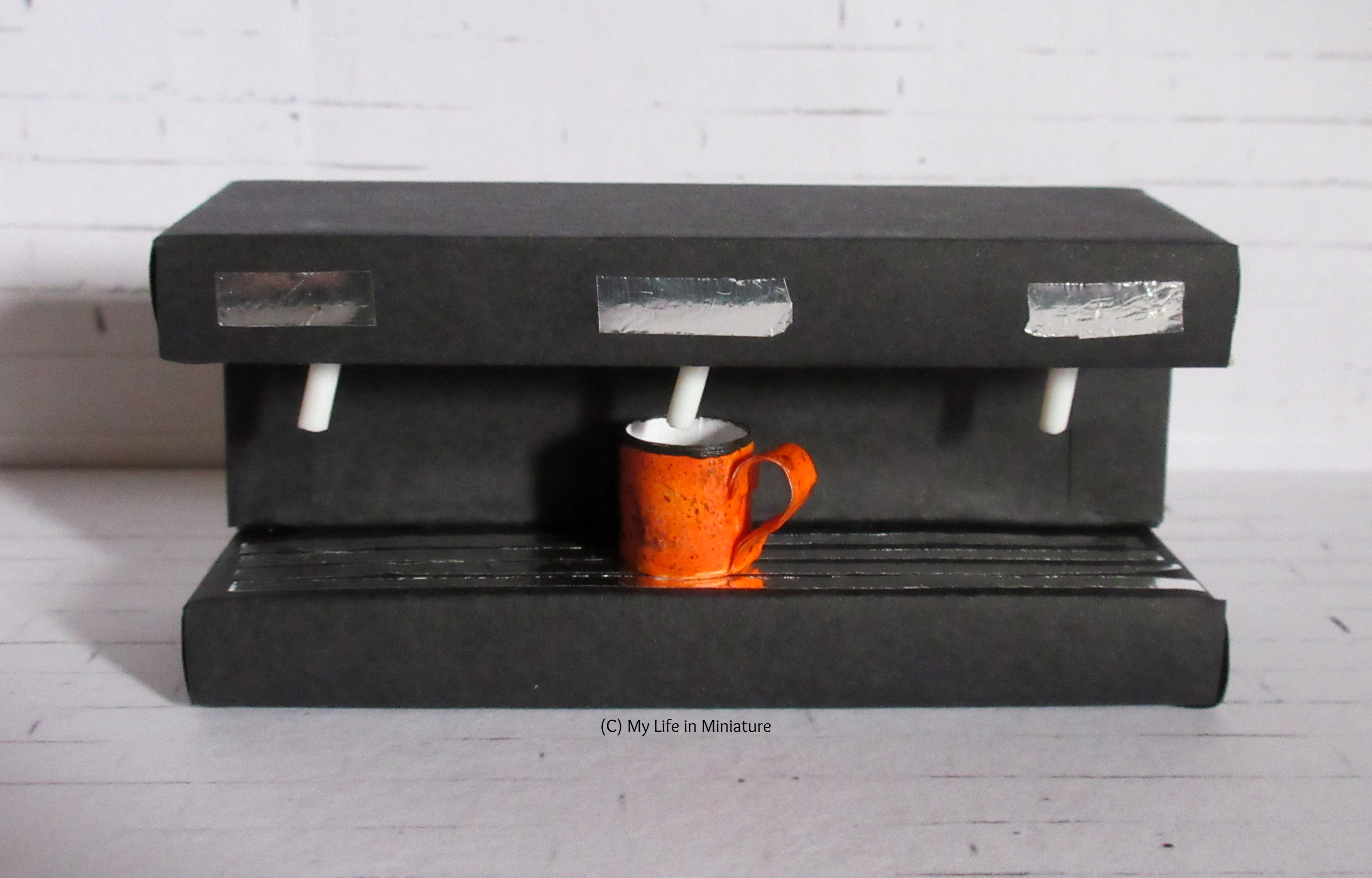 A large rectangular coffee making-machine sits against a white brick background. There are three spots for cups to go under, and an orange tin mug is under the middle one.