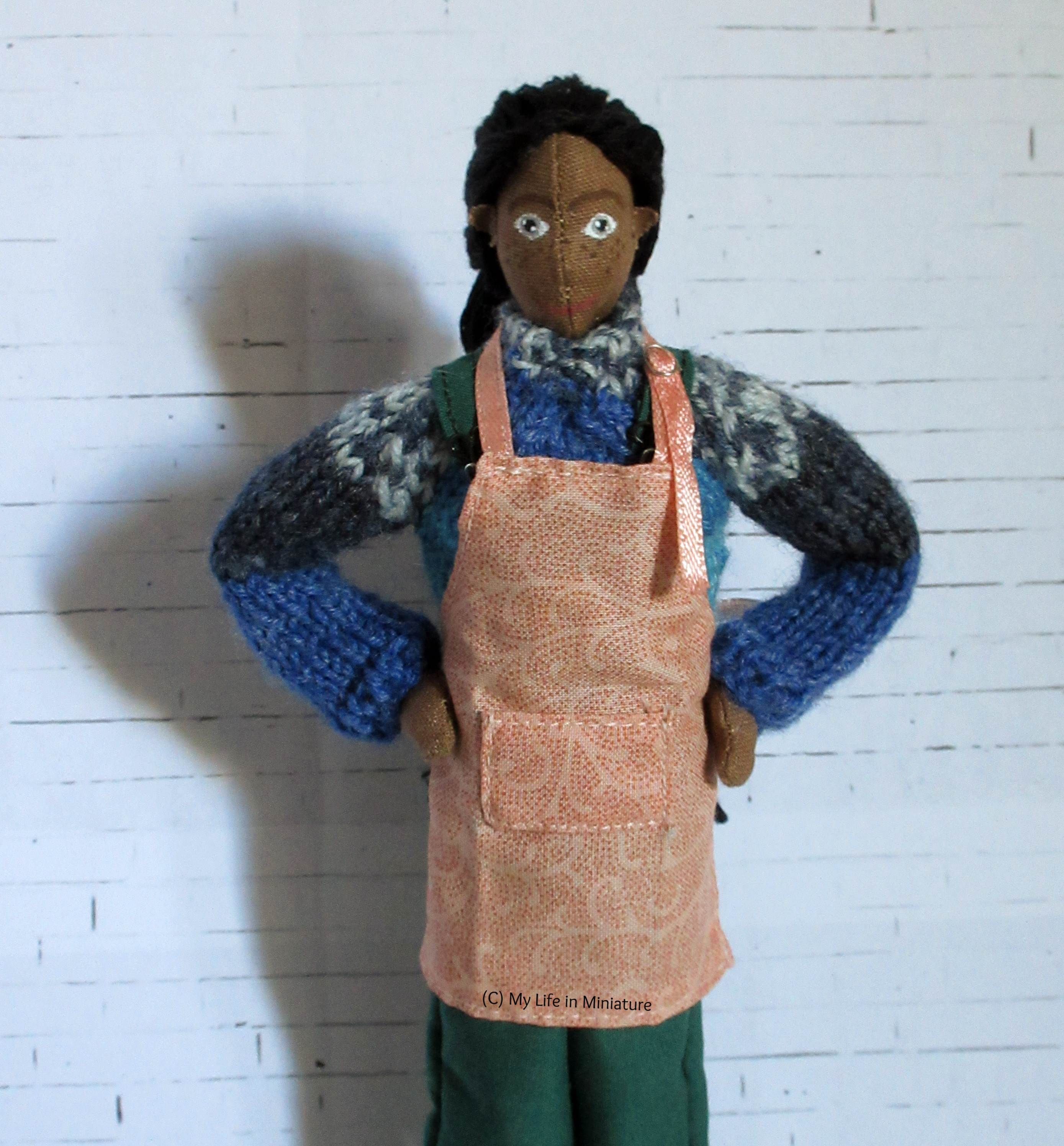 Petra stands against a white brick background, hands on her hips and looking at the camera. She wears a pale pink apron with a front pocket and a sliding buckle on the neck strap. 