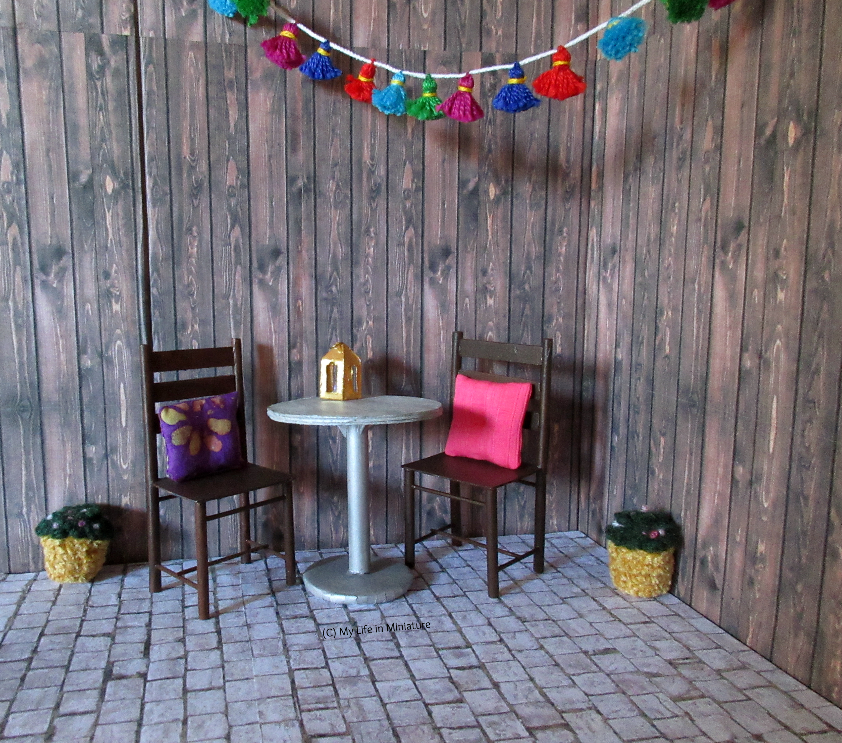 The courtyard outside Tea at Yaz's is pictured. It has dark wood walls and cobblestones, and is furnished with two brown chairs and a silver circular table. Two small flower pots are also on the floor, and a garland of multi-coloured tassels hangs across the top.