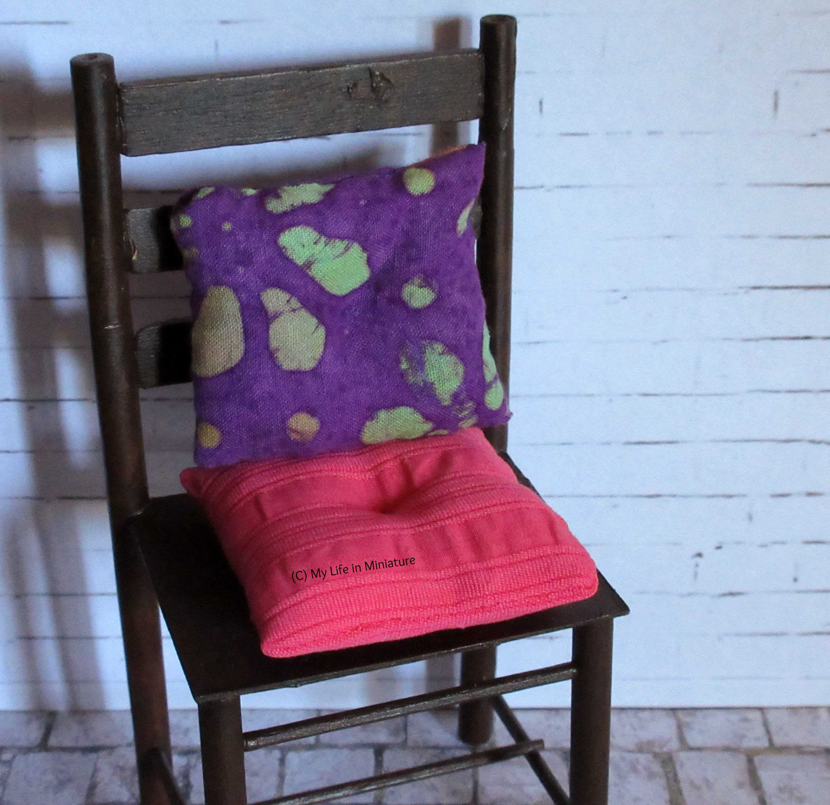 One of the brown chairs sits against a white brick background. On it are two cushions: one in purple batik fabric, one in pink fabric. Both have divots sewn in the centre. 