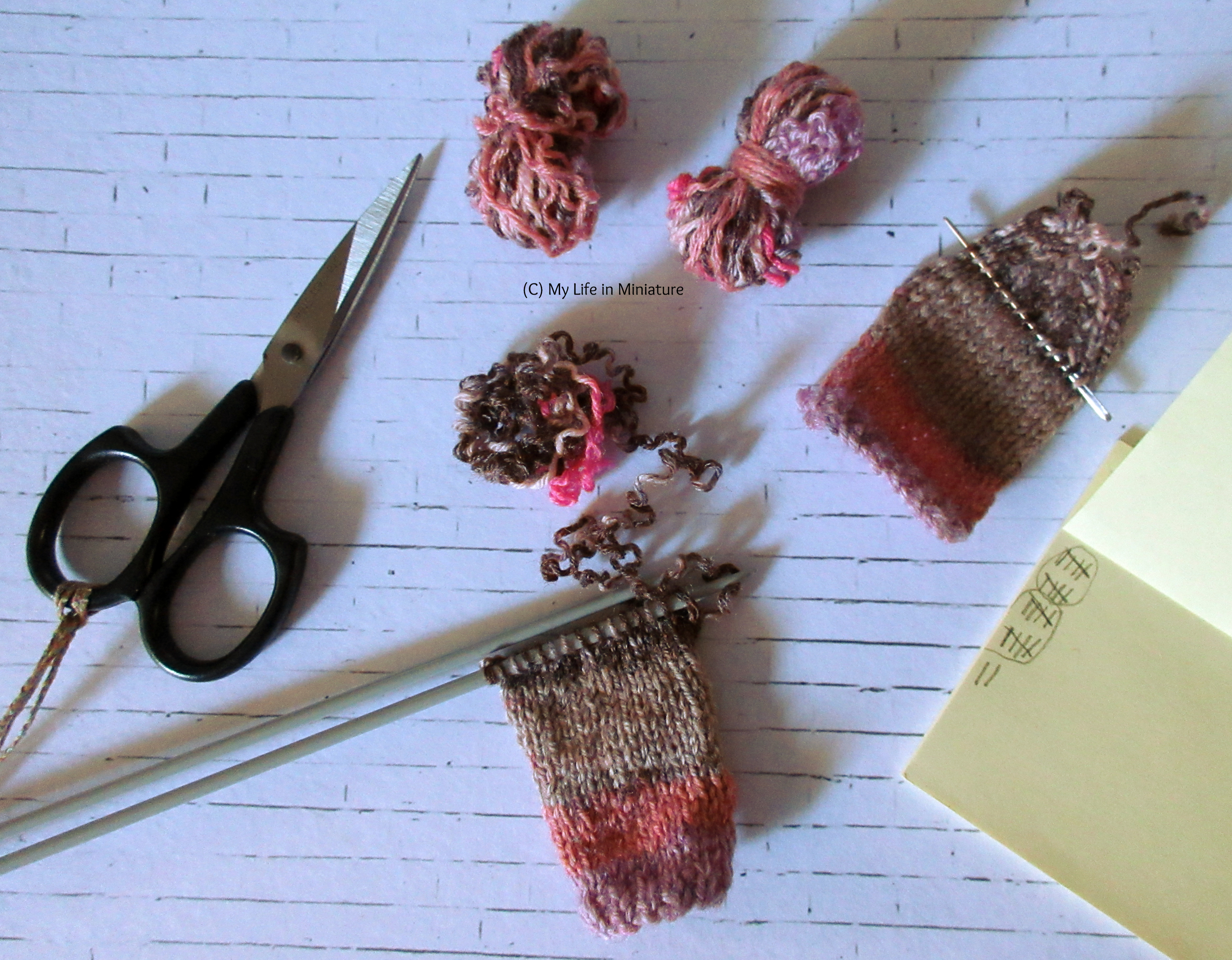 The pink jumper is in pieces on a white brick background. One of the sleeves is being re-knitted, and the front and back panels are two small skeins at the top of the image.