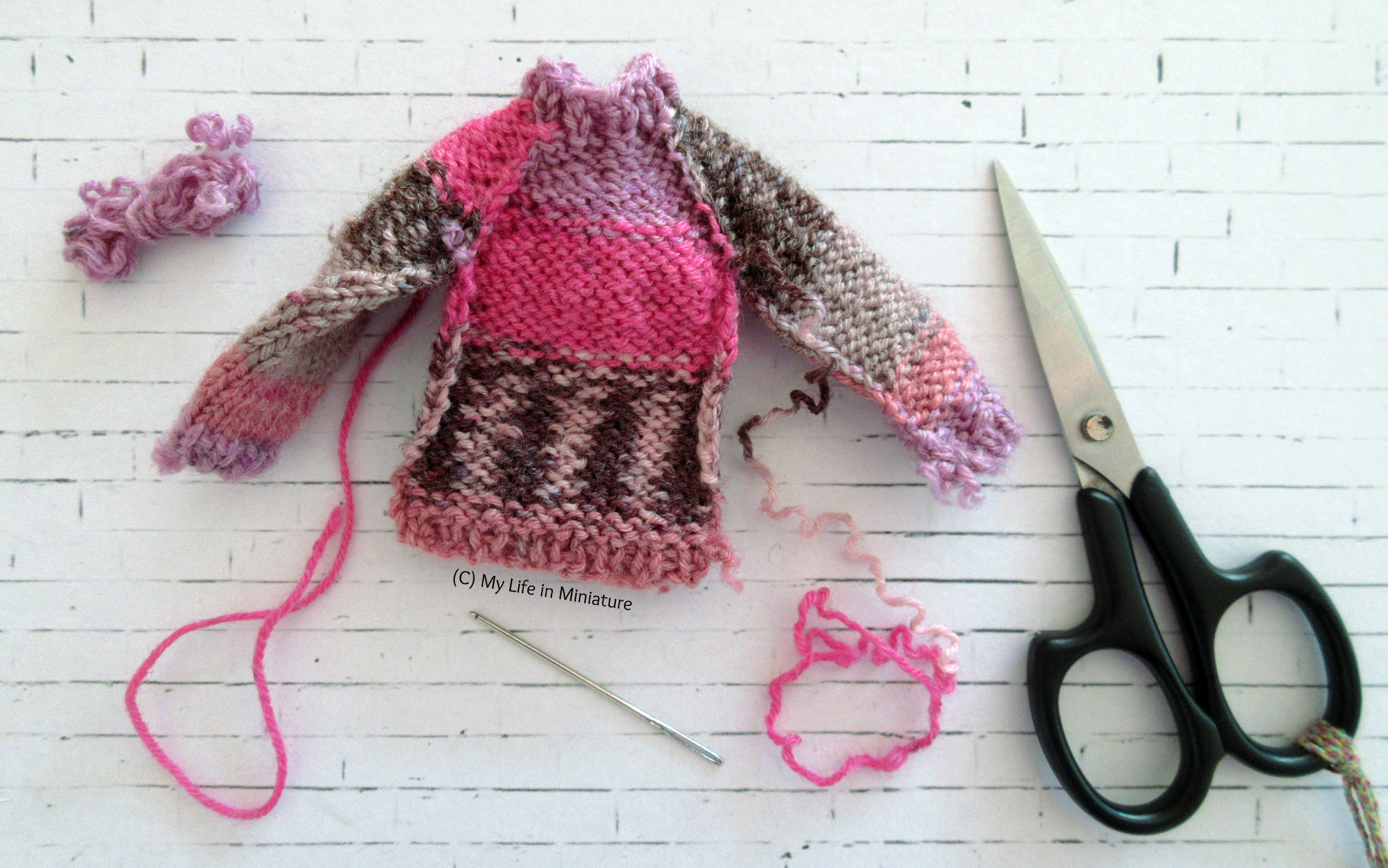 The pink jumper is shown mid-assembly on a white brick background. The four panels are sewn together, but the arm and side seams are not sewn yet. A needle and a pair of embroidery scissors are also visible.