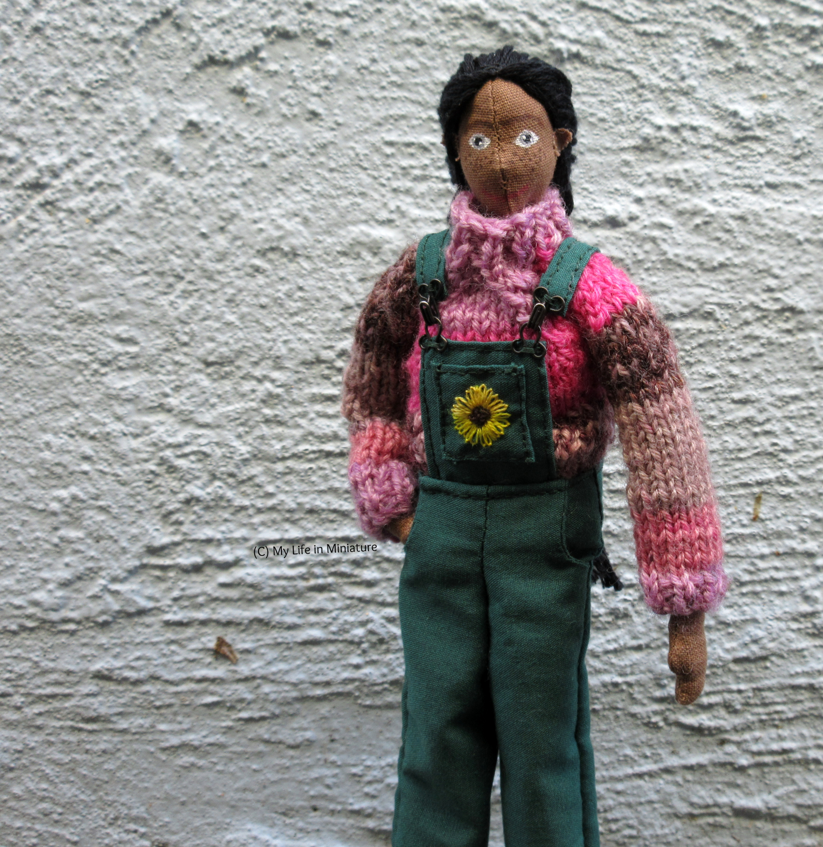 Petra wears the pink jumper under her green dungarees, one hand in her pocket. She's outside, against a pale blue textured wall, looking at the camera.