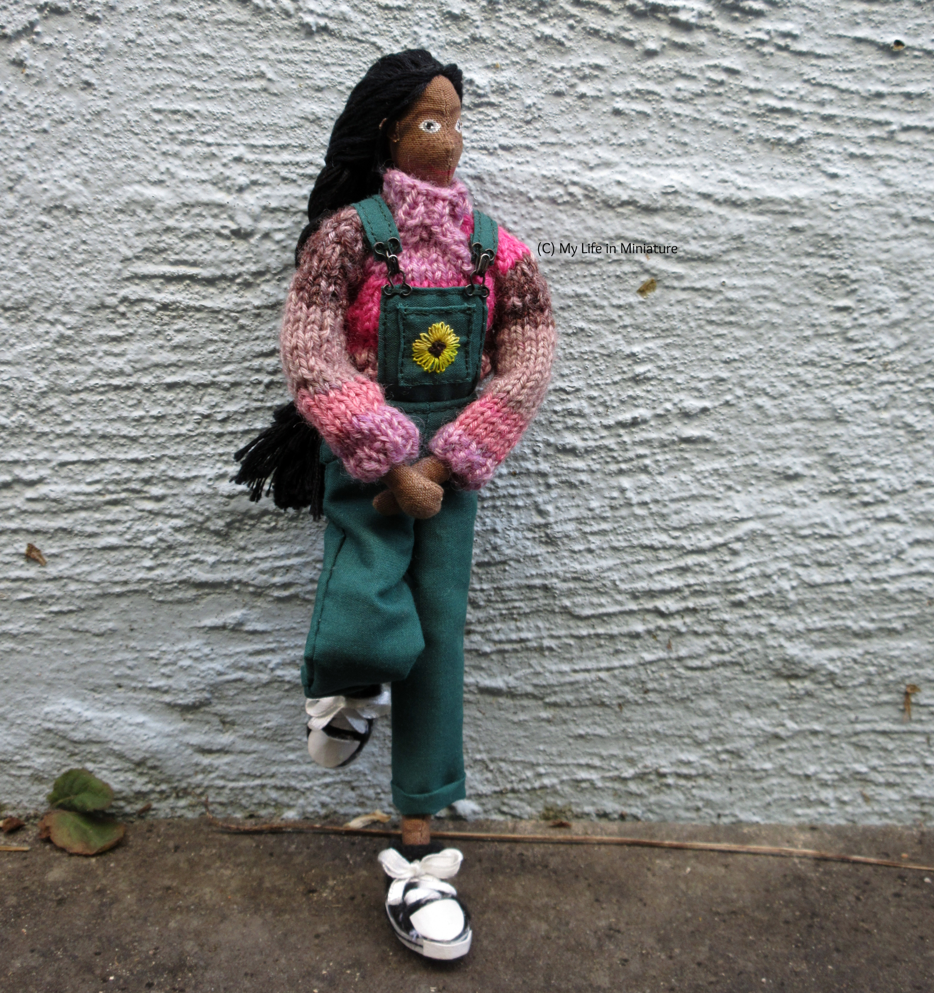 Petra wears the pink jumper, green dungarees, and black shoes against an outdoor textured wall. She has one foot up on the wall, and her hands in front of her. She looks to the right.
