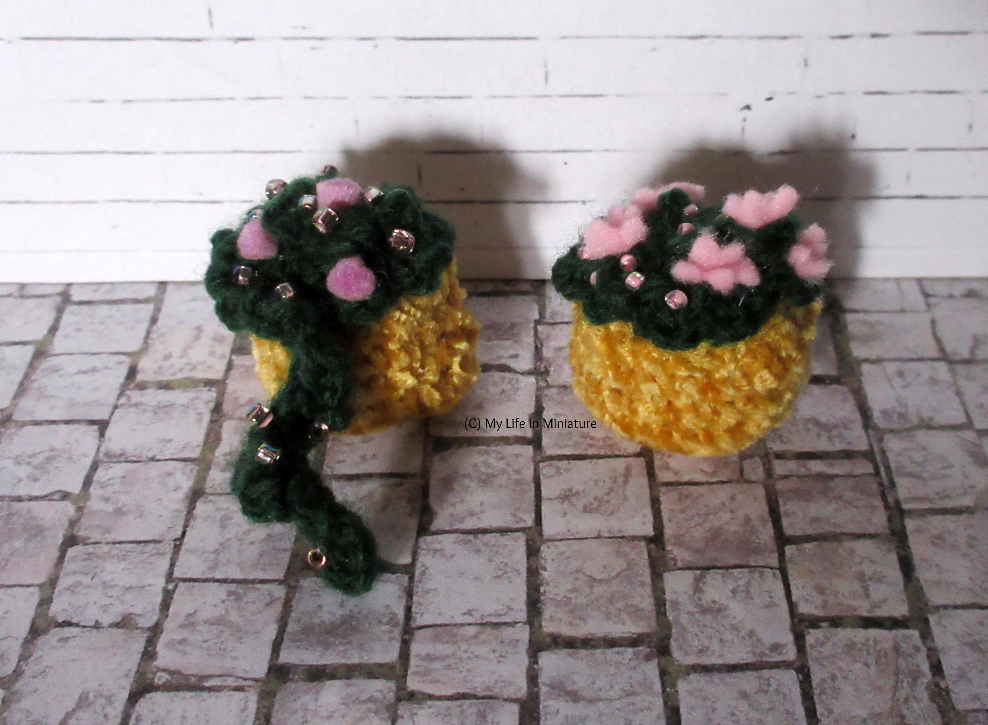 Two of the baskets are against a white brick background. One has purple flowers and beads in, with one stem (?) dangling over. The other has larger pink flowers and beads in. The pots are yellow.