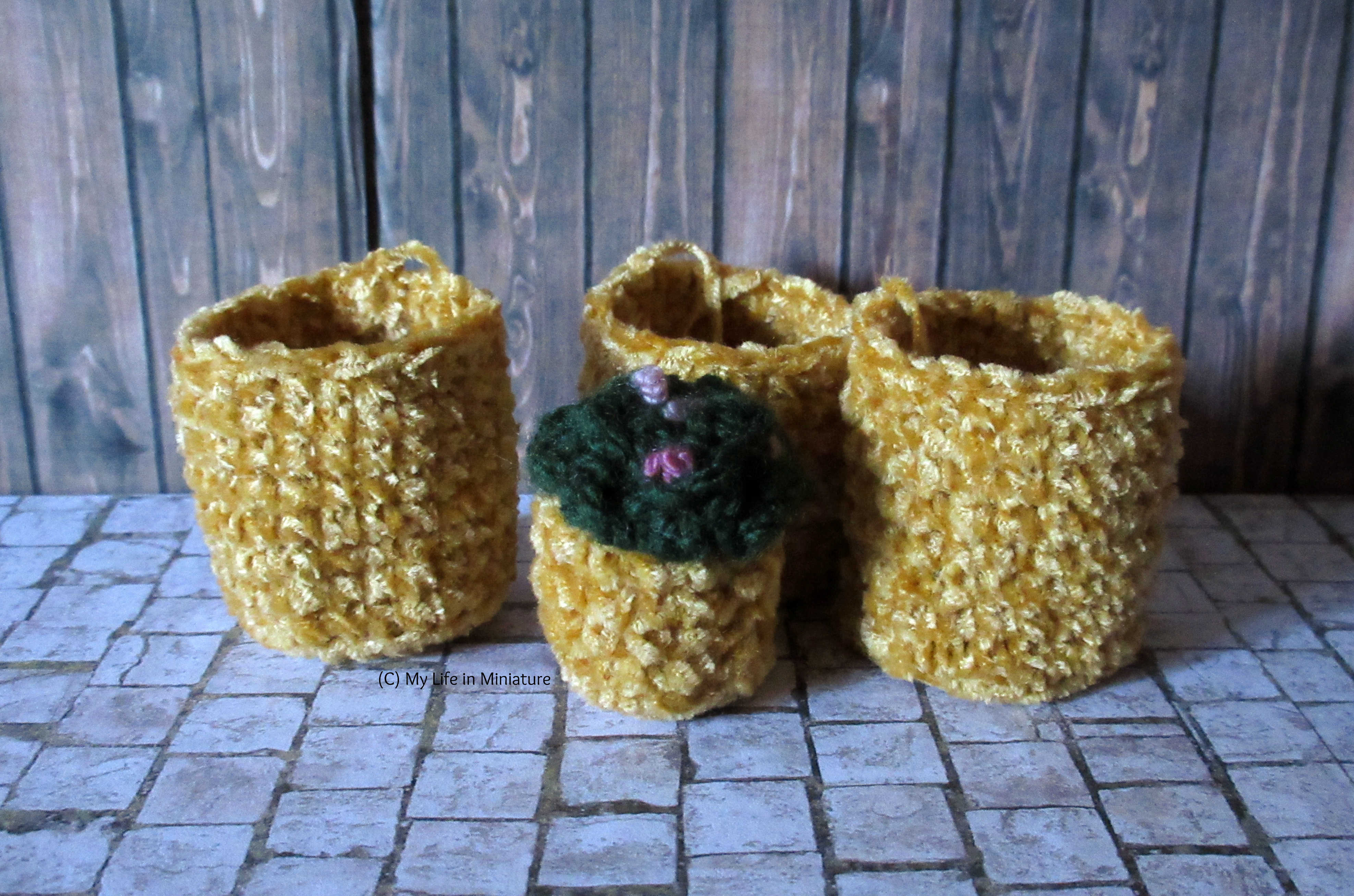 Three large crocheted pots stand behind one of the smaller pots in Tea at Yaz's courtyard. The big pots are the height of the smaller pot, including the greenery.