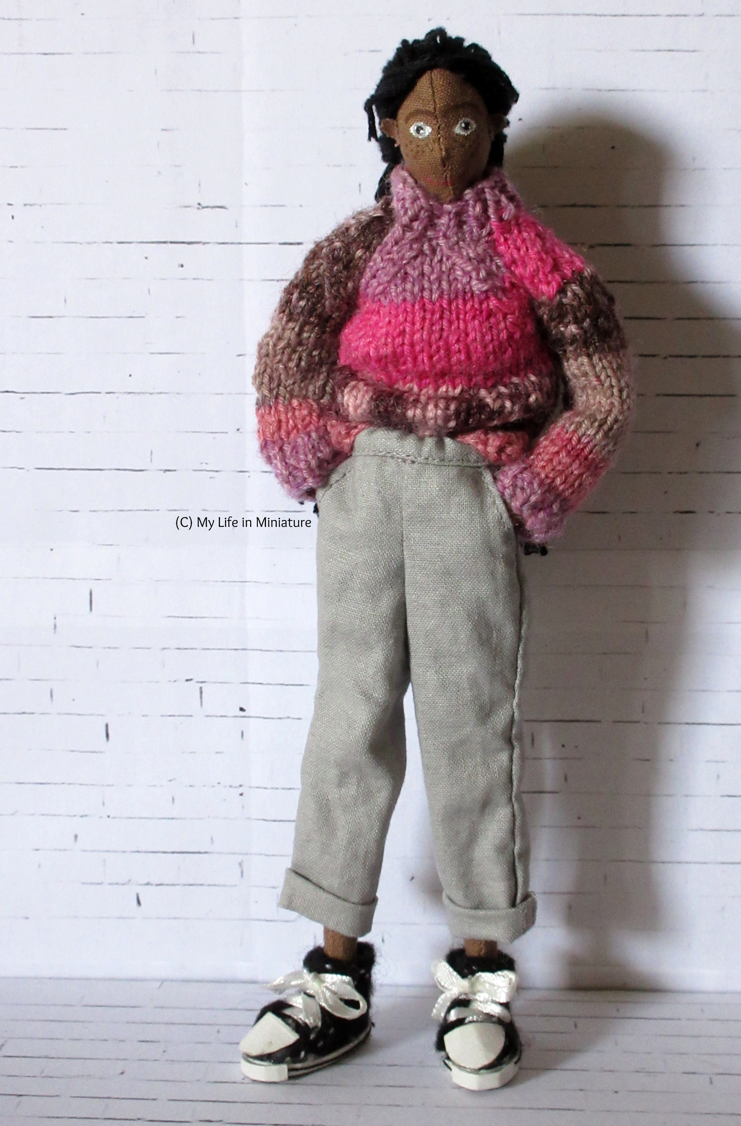 Petra stands against a white brick background. She wears loose pale grey pants, rolled up at the hem, with her pink jumper. Her hands are in her pockets, and she looks at the camera. 