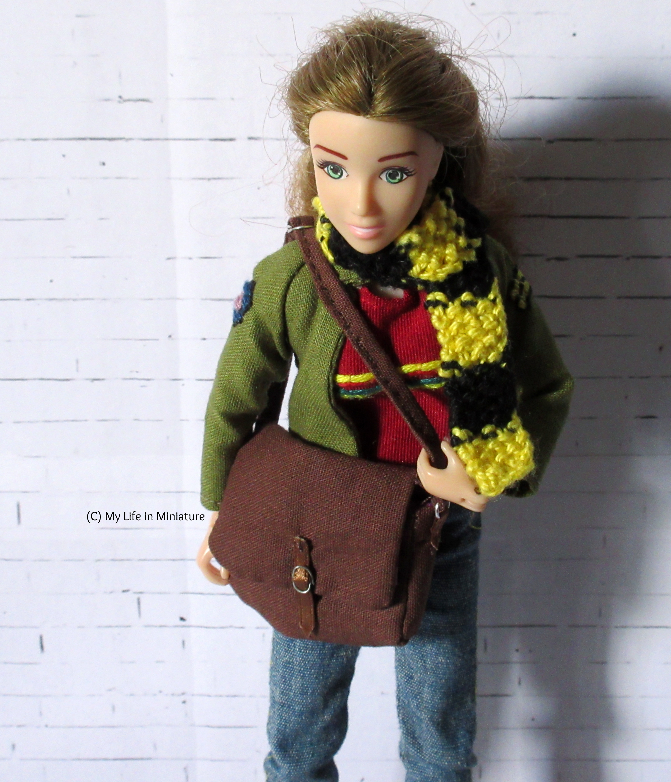 Sarah stands in front of a white brick background. She has a brown fabric bag over her shoulder, which she's pulling around to the front for the camera. She's looking down at the bag. 