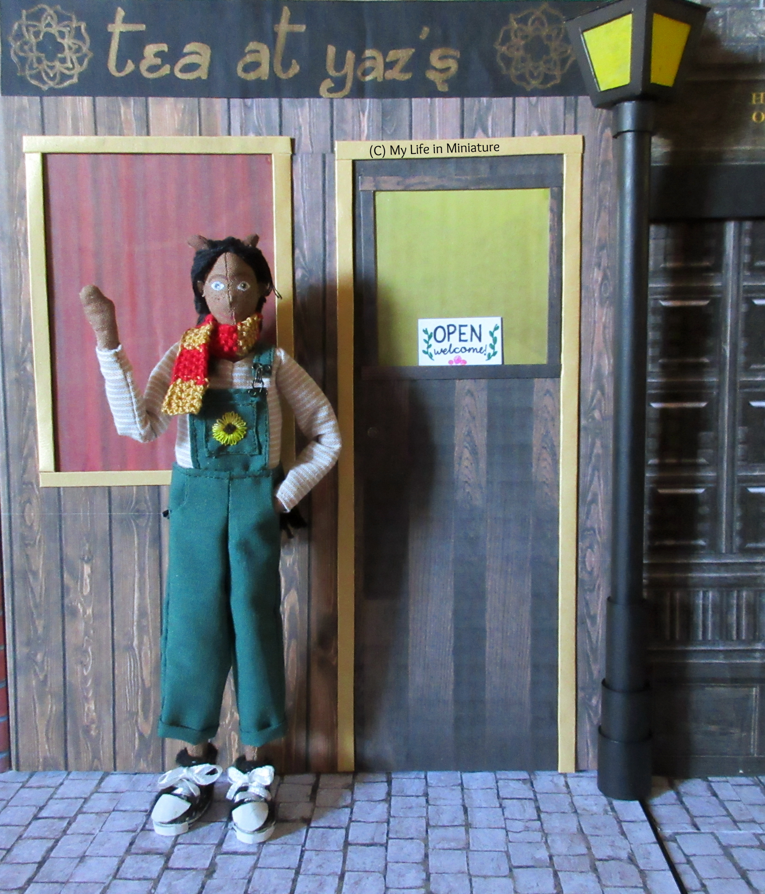 Petra stands outside Tea at Yaz's, which has an 'Open; welcome!' sign on the door. She waves at the camera. 